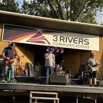 Group of people performing on an outdoor stage with a banner reading '3 Rivers Communications'.