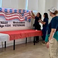 People hanging patriotic decorations reading 'FALLS Welcome VETERANS' in a community center.