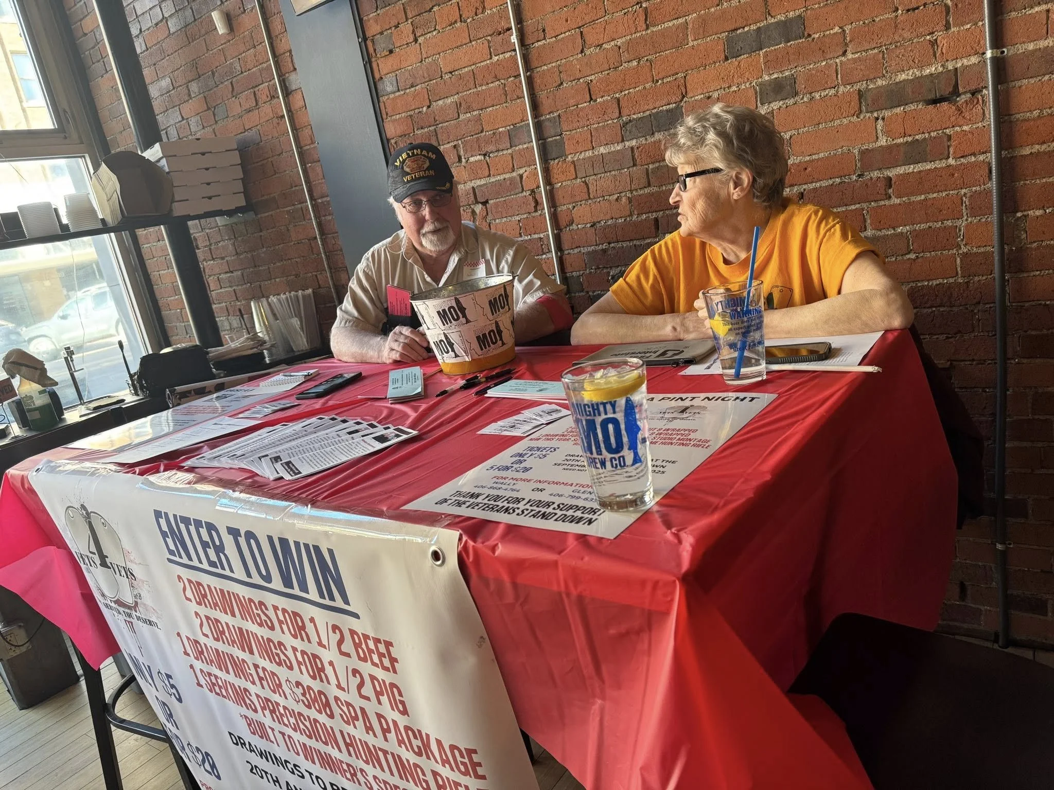 A man wearing a Vietnam veteran cap and a woman with glasses sitting at a table covered with a red tablecloth, engaging in conversation. The table has various papers, drinks, and a large promotional banner about a contest for drawing and precision hunting.