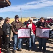 Group of five people standing outdoors holding signs, with cars parked in the background under a partly cloudy sky.