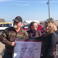 Three people standing outdoors, holding a large check, with cars and a utility pole in the background.