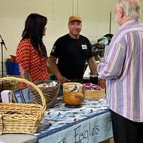 A vendor at a market stall speaking with two customers, with baskets and baked goods displayed on the table.