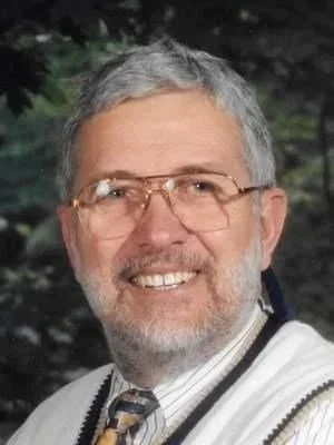 A middle-aged man with gray hair and beard, wearing glasses, a patterned tie, and a sweater, smiling outdoors with trees in the background.