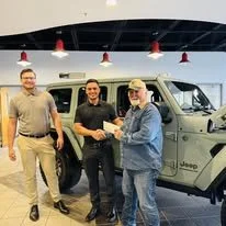 Three men standing inside a car dealership, shaking hands in front of a new beige Jeep.