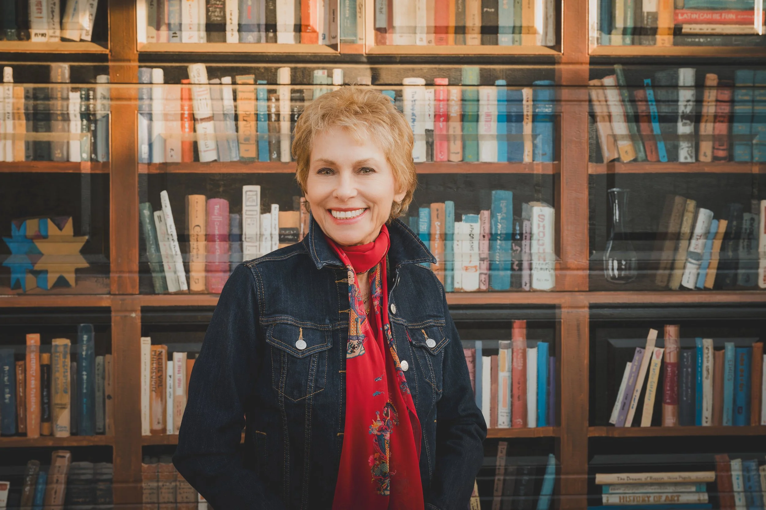 Merry Elkins with short blonde hair wearing a denim jacket and red scarf, standing in front of a bookshelf filled with books.