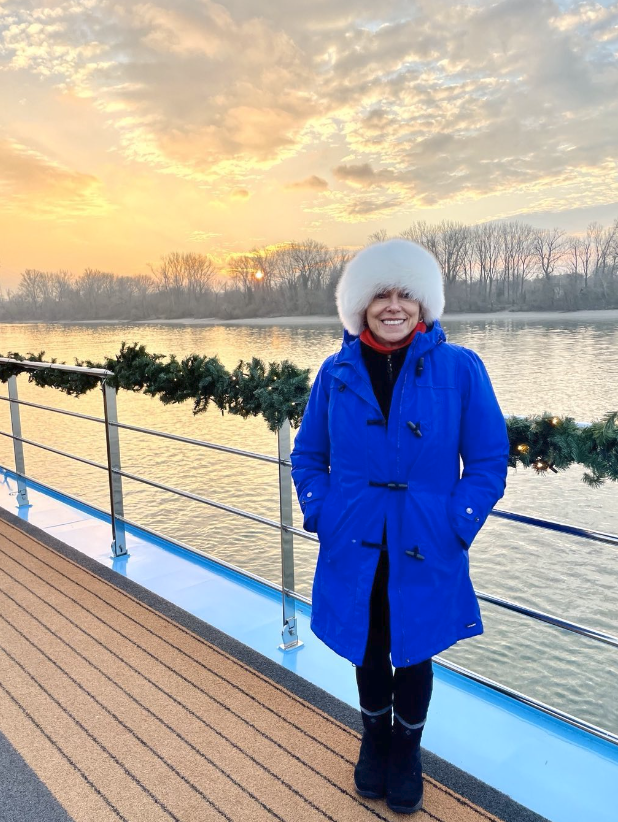 Merry Elkins wearing a white furry hat and a bright blue winter coat standing on a boat deck with a river and leafless trees in the background during sunset.