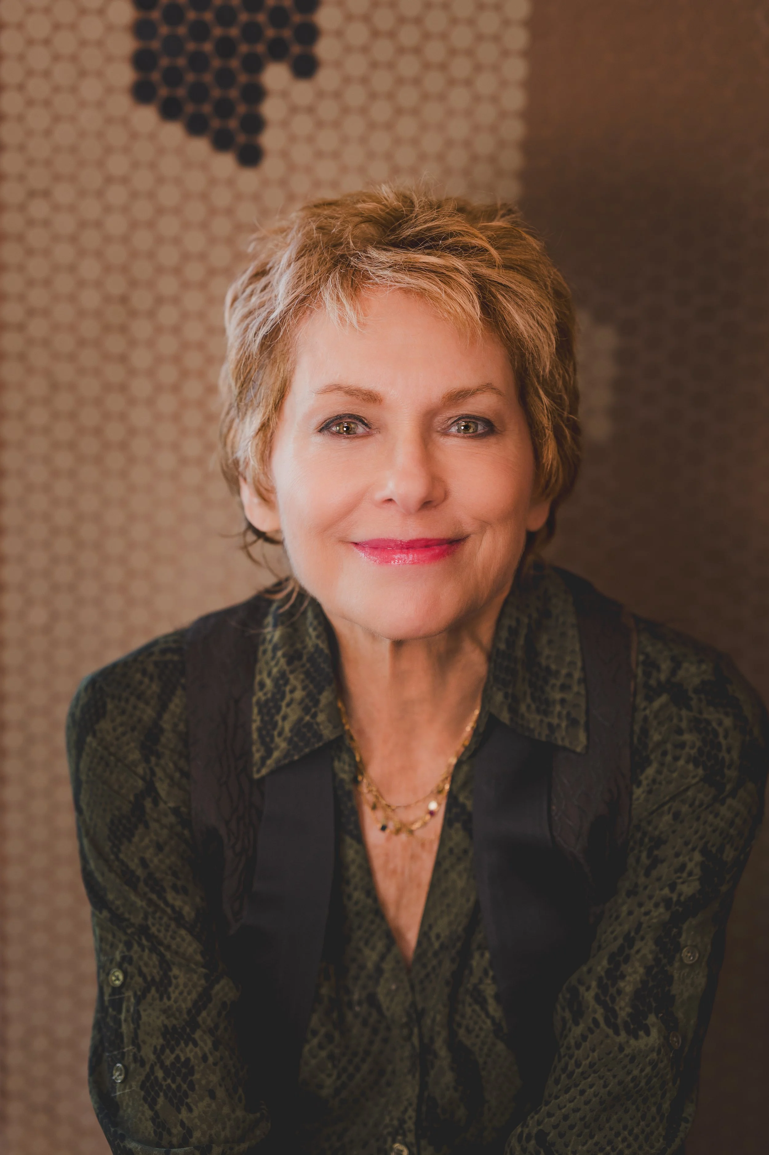Writer, Merry Elkins with short, layered blonde hair, smiling, wearing a dark green patterned blouse and a gold necklace, standing in front of a textured wall with black and beige hexagonal patterns.