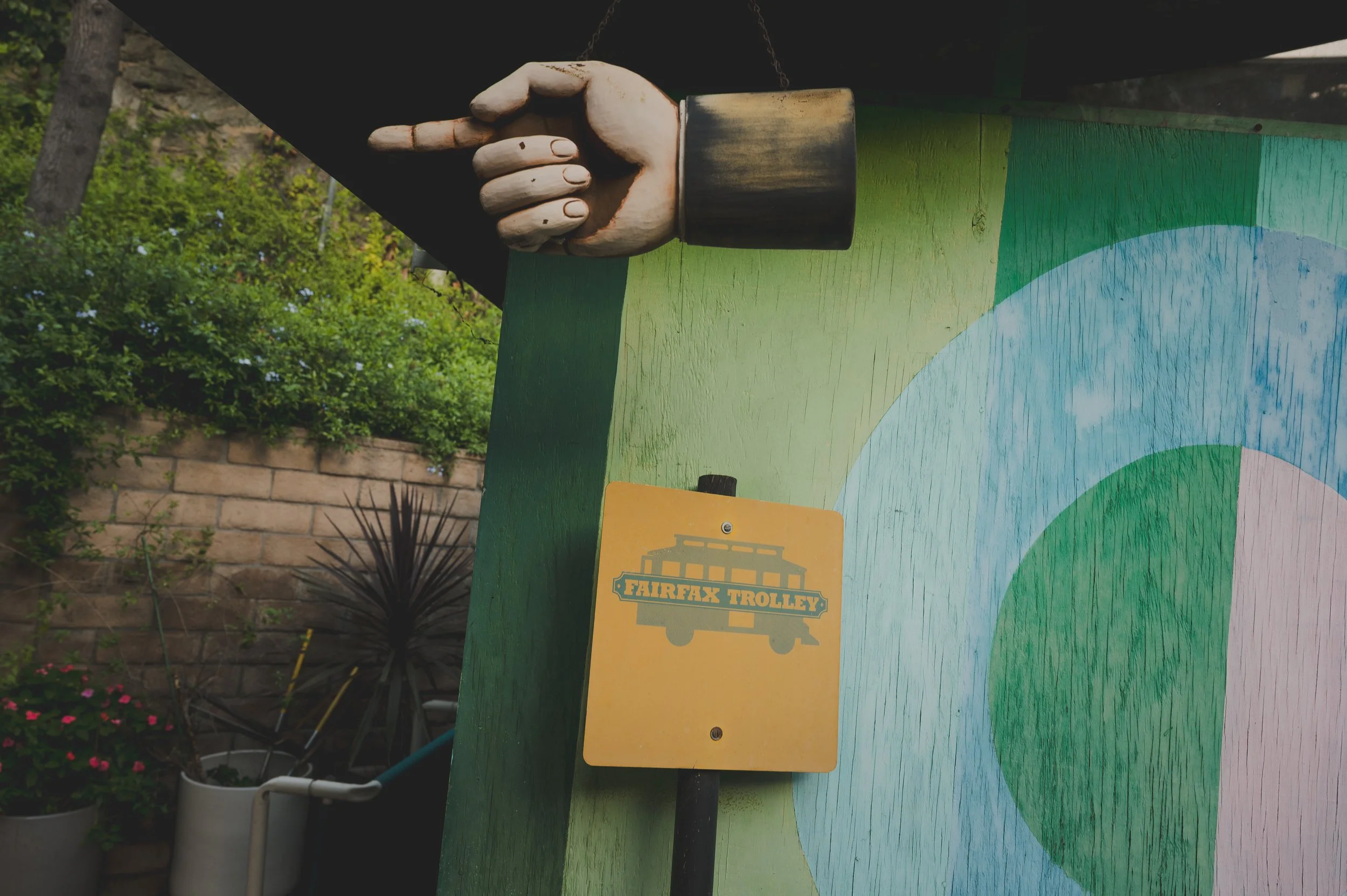 A painted mural featuring a large hand with a painted wooden hand pointing with the index finger, above a yellow sign with a trolley icon and the words 'Fairfax Trolley', set against a colorful background with green, blue, and pink sections.