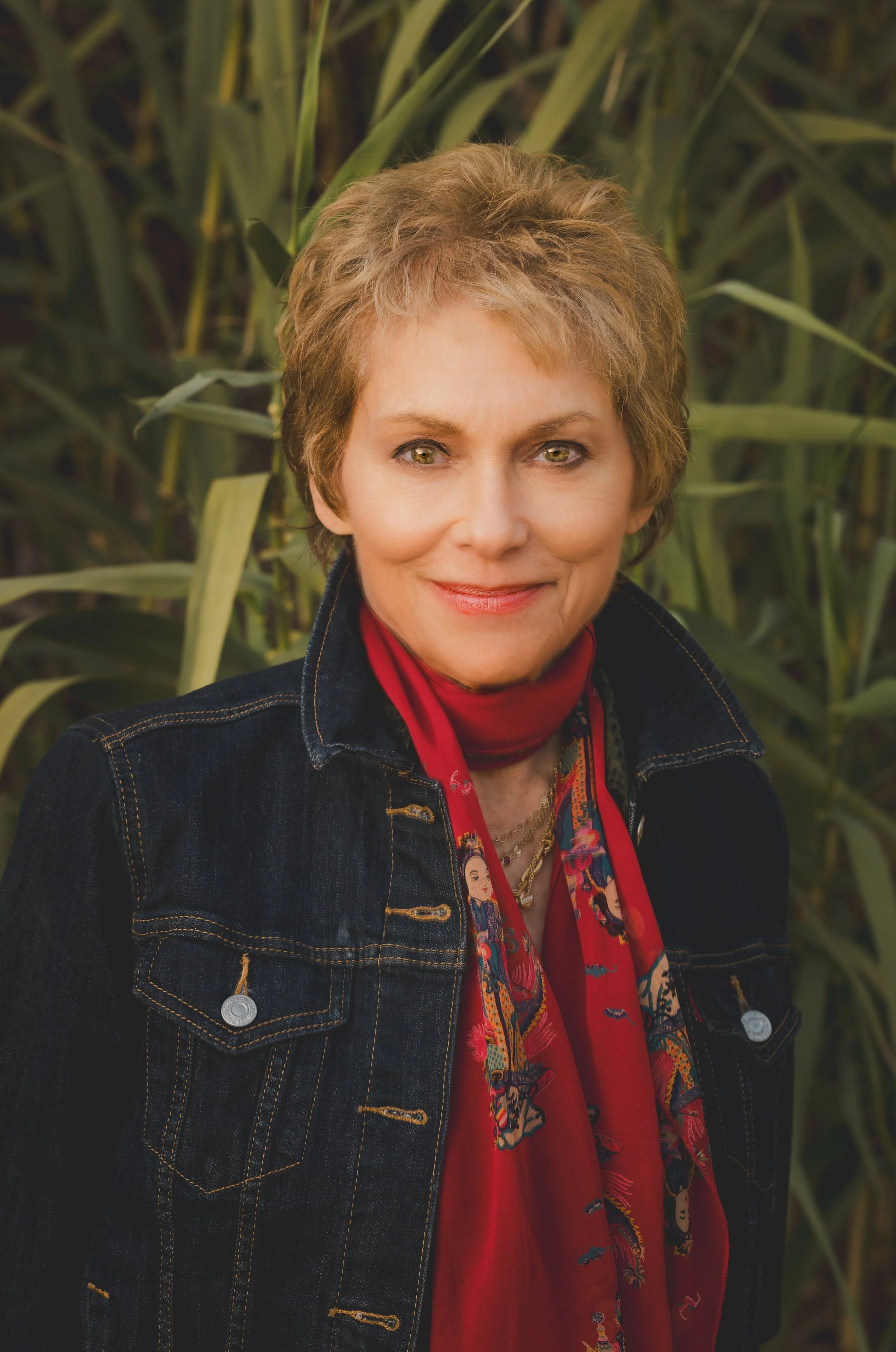 Writer, Merry Elkins with short light brown hair, yellow eyes, and a friendly smile standing in front of tall green plants, wearing a denim jacket and a red scarf.