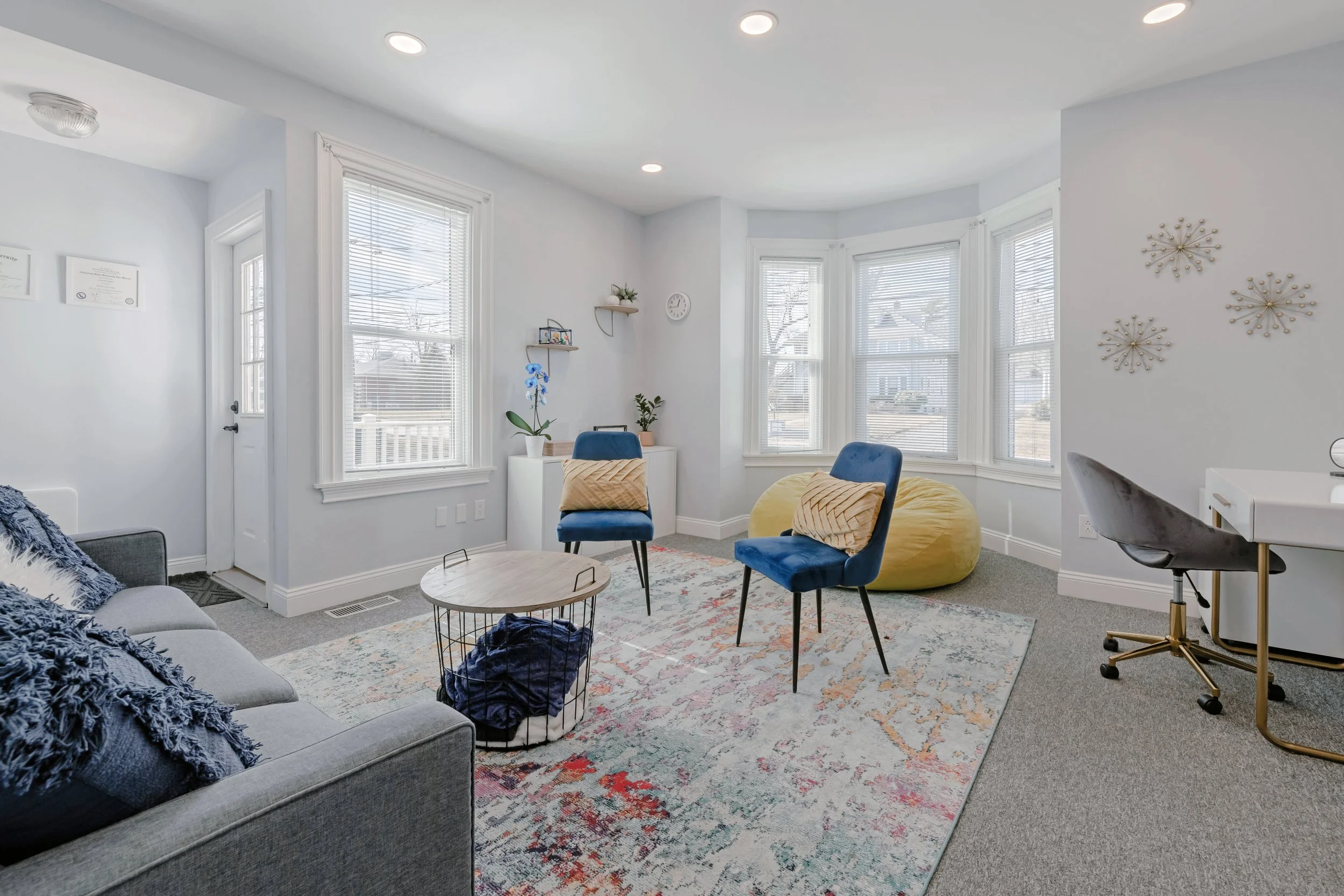 Room with gray sofa, blue chairs, yellow bean bag, patterned area rug, and multiple windows with white blinds.
