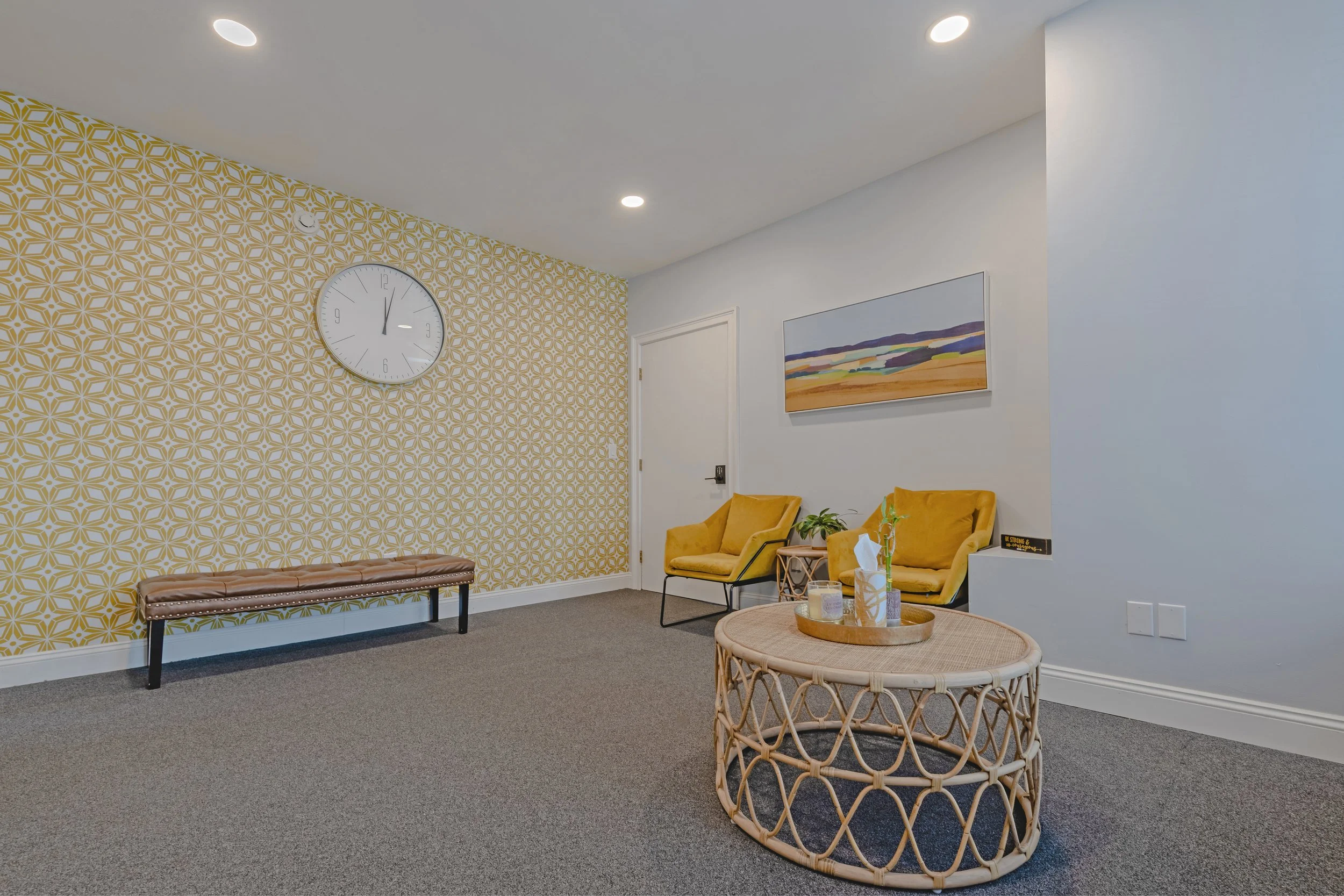 A modern waiting room with yellow patterned accent wall, two yellow armchairs, a rattan coffee table with decorative items, a brown bench, a large round wall clock, and a colorful landscape painting.