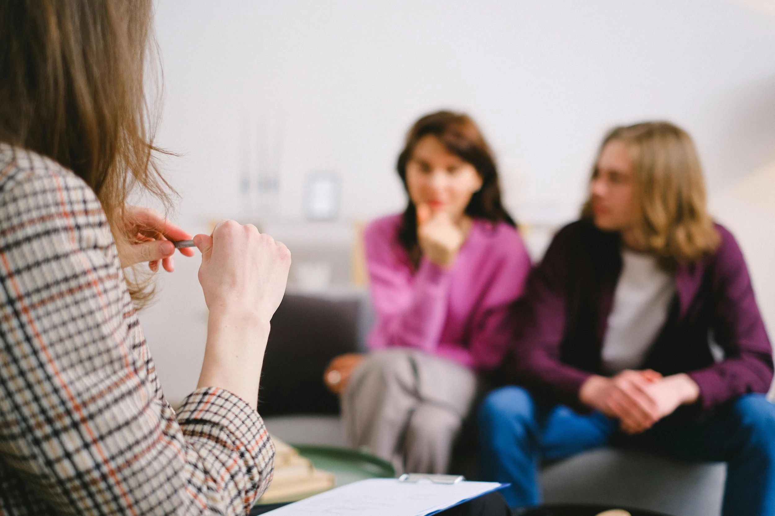 A woman taking notes during a therapy or counseling session with two women who are seated and listening attentively.