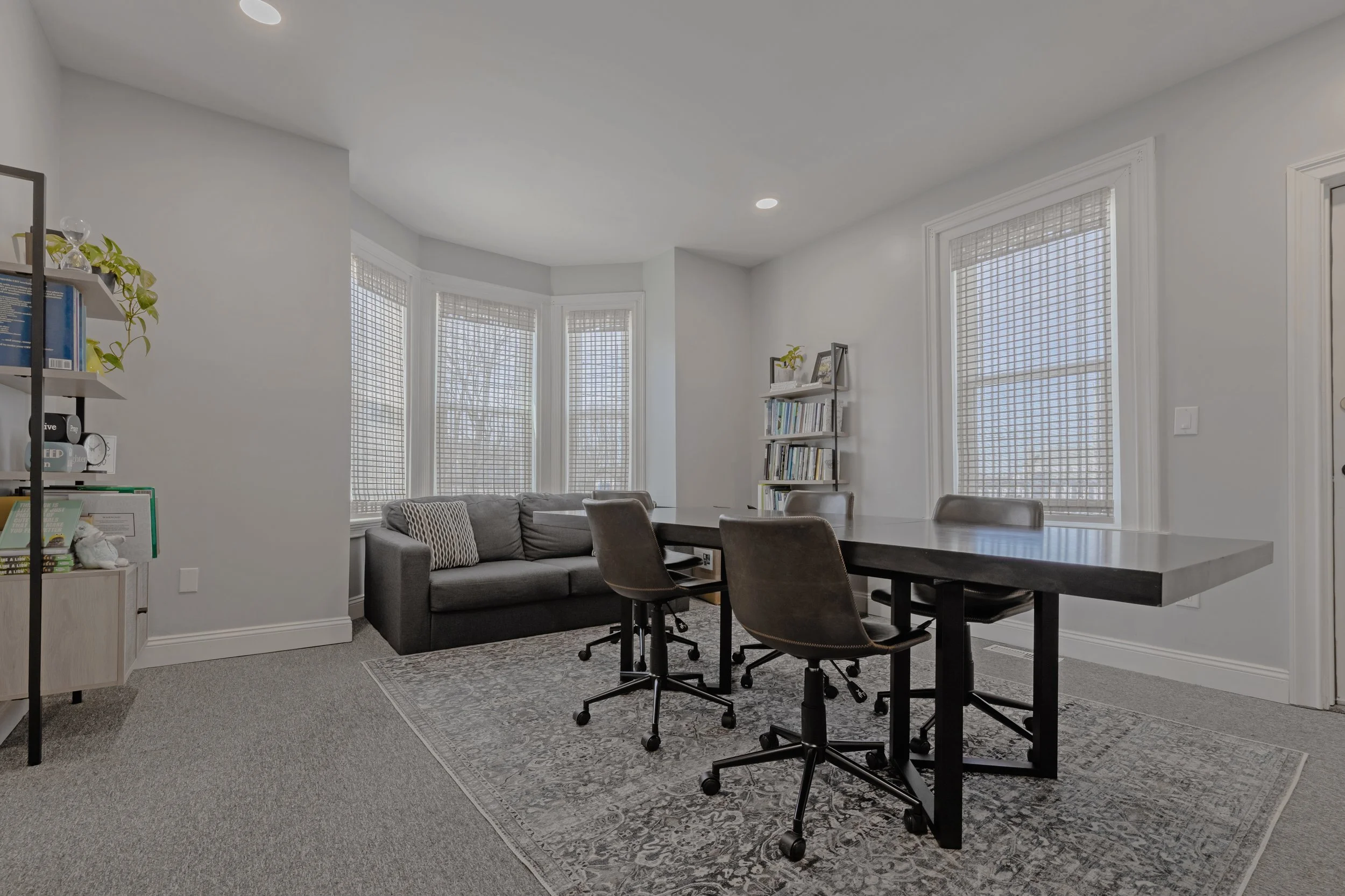 Room with a dark wooden dining table, four office chairs on wheels, a gray sofa, bookshelves, and large windows with blinds.