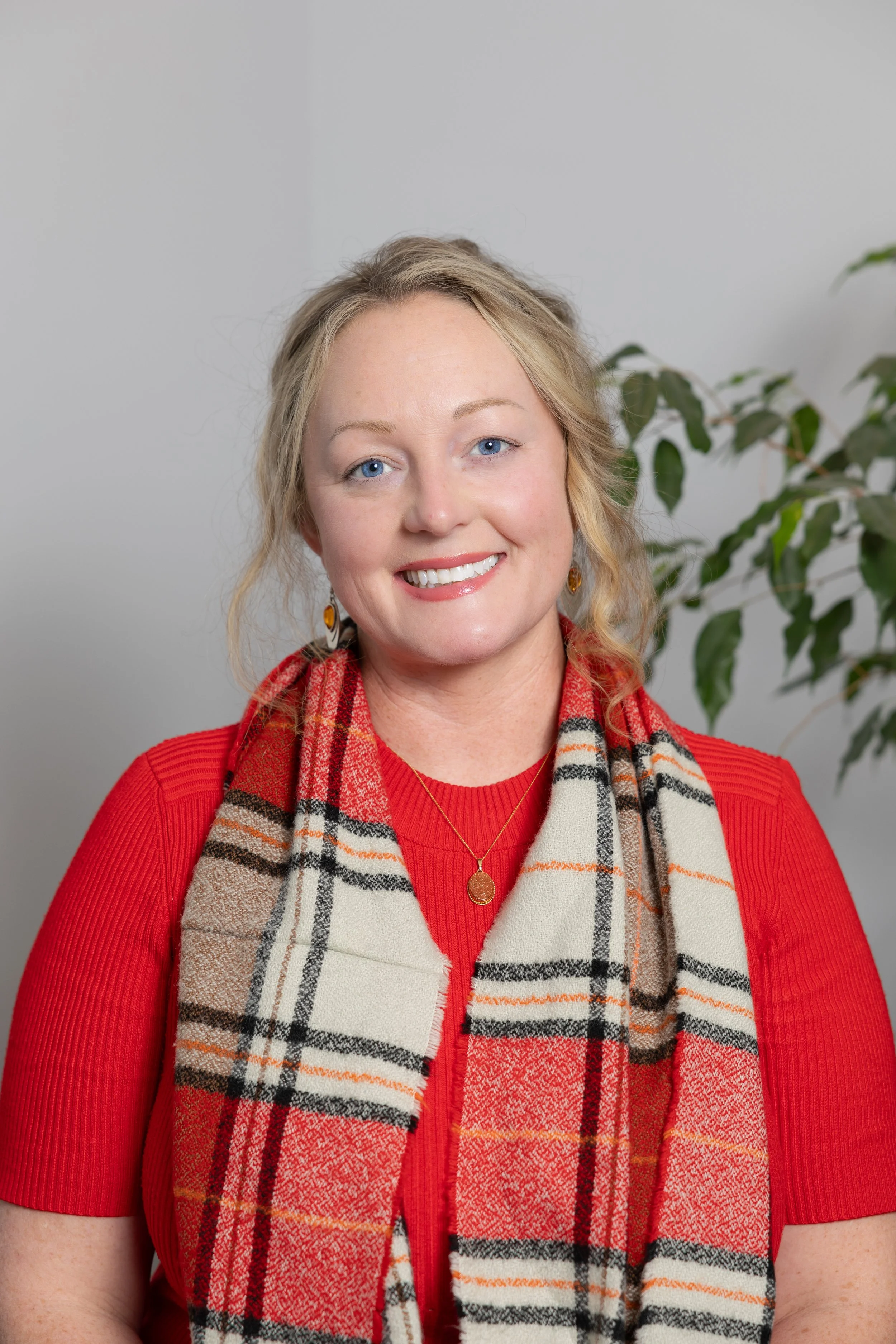 A woman with blonde hair, blue eyes, smiling, wearing a red top, a plaid scarf, a necklace, and earrings, in front of a neutral wall with green plant.