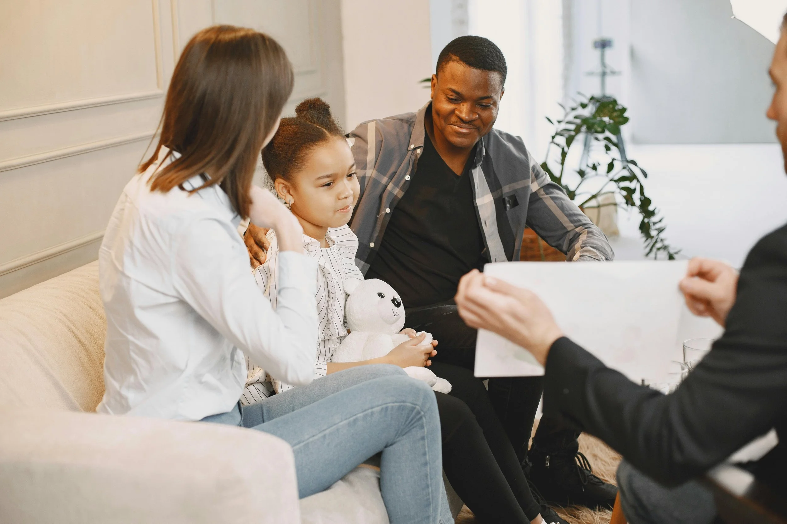A young girl holding a stuffed animal, sitting on a couch with a woman and a man, while a person in black documents something on paper.