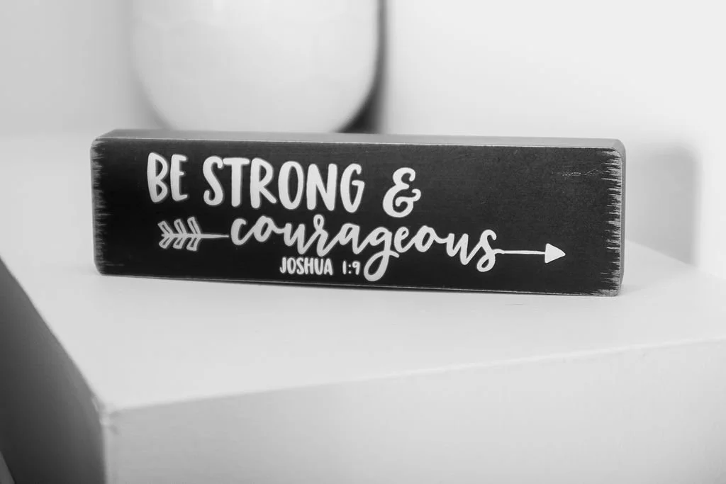 A black rectangular wooden plaque with white handwritten-style text that says "Be strong & courageous" and includes a reference to Joshua 1:9, placed on a white surface with a blurred background.