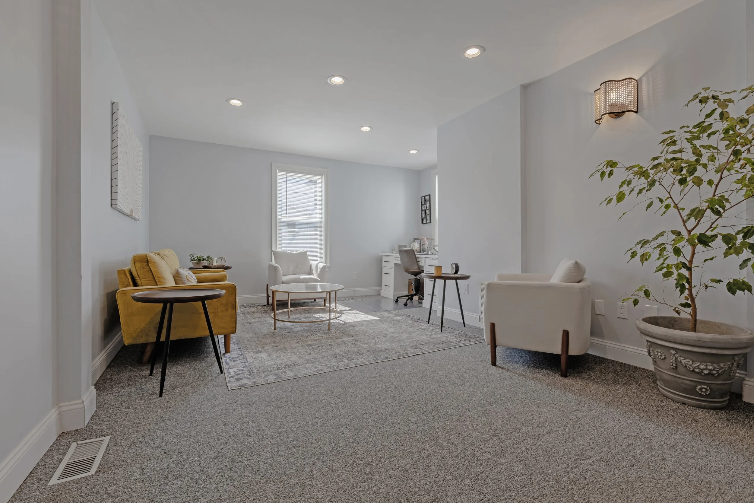 Room with white walls, recessed ceiling lights, a large window, a yellow sofa, white armchairs, a rug, small tables, a desk with a chair, and a potted plant.