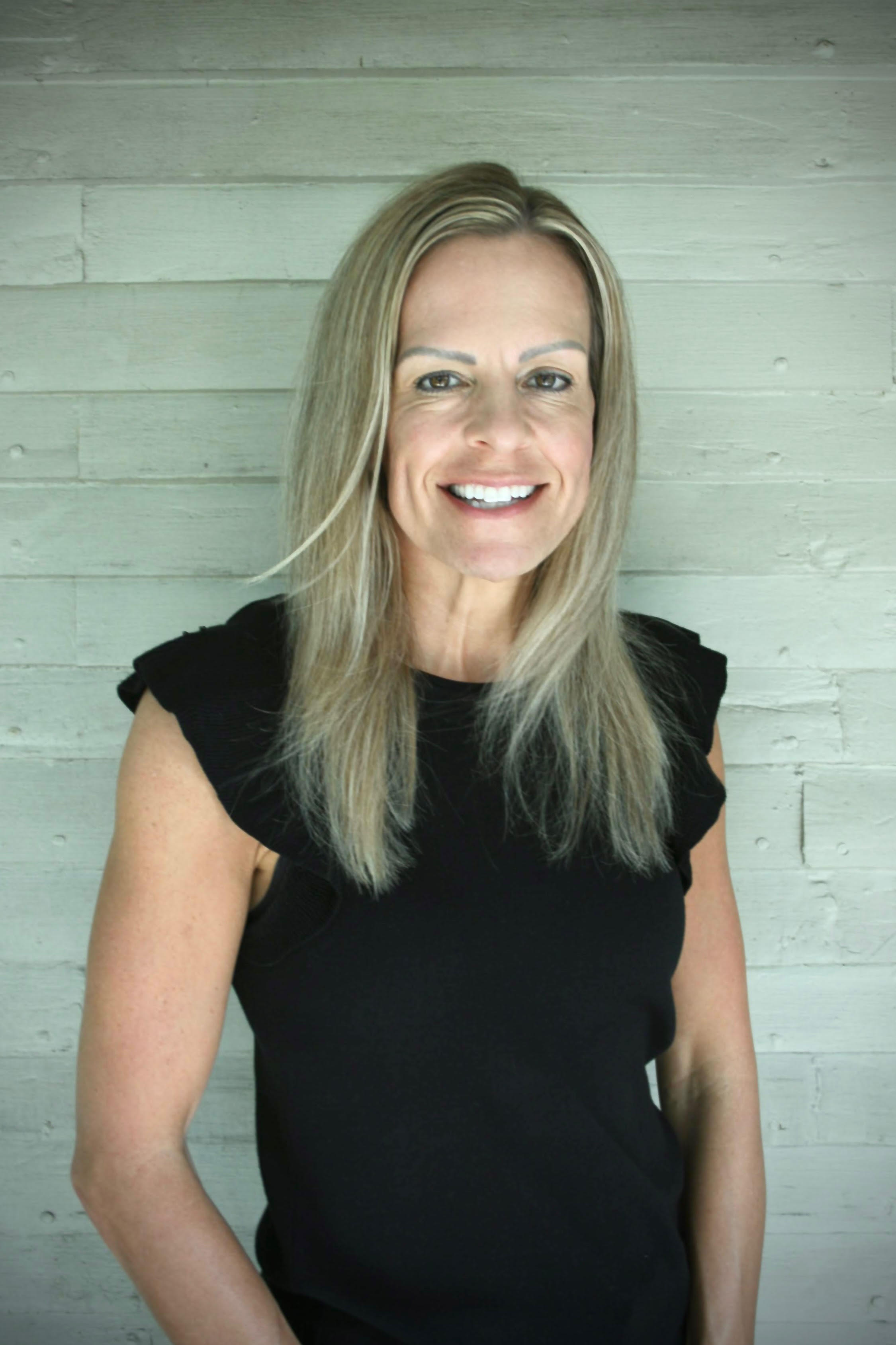 A woman with shoulder-length blond hair, smiling, wearing a black sleeveless top, standing against a light gray wooden wall.