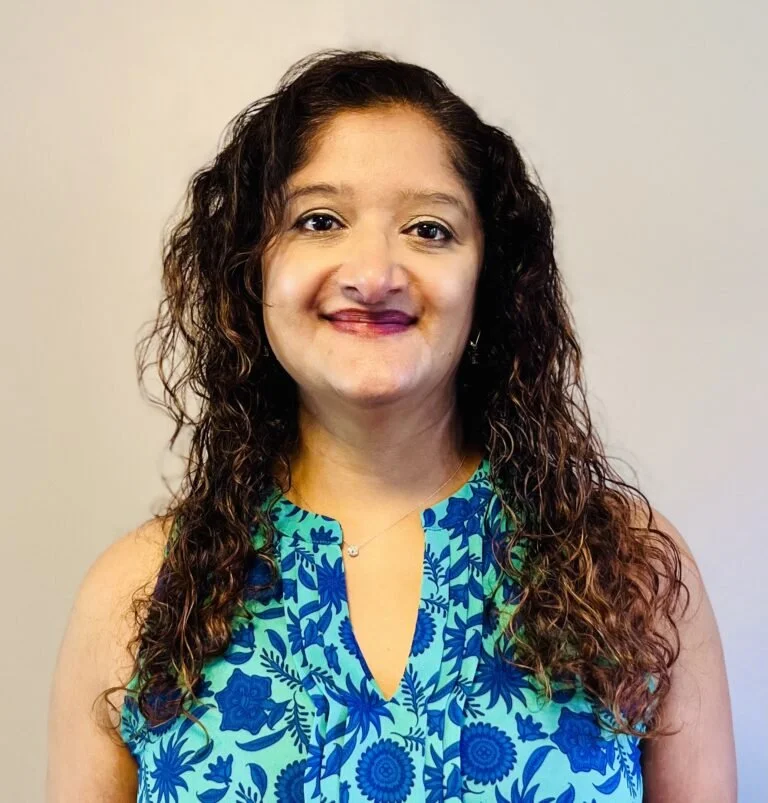 A woman with curly brown hair wearing a sleeveless blue floral top standing against a plain light gray wall.