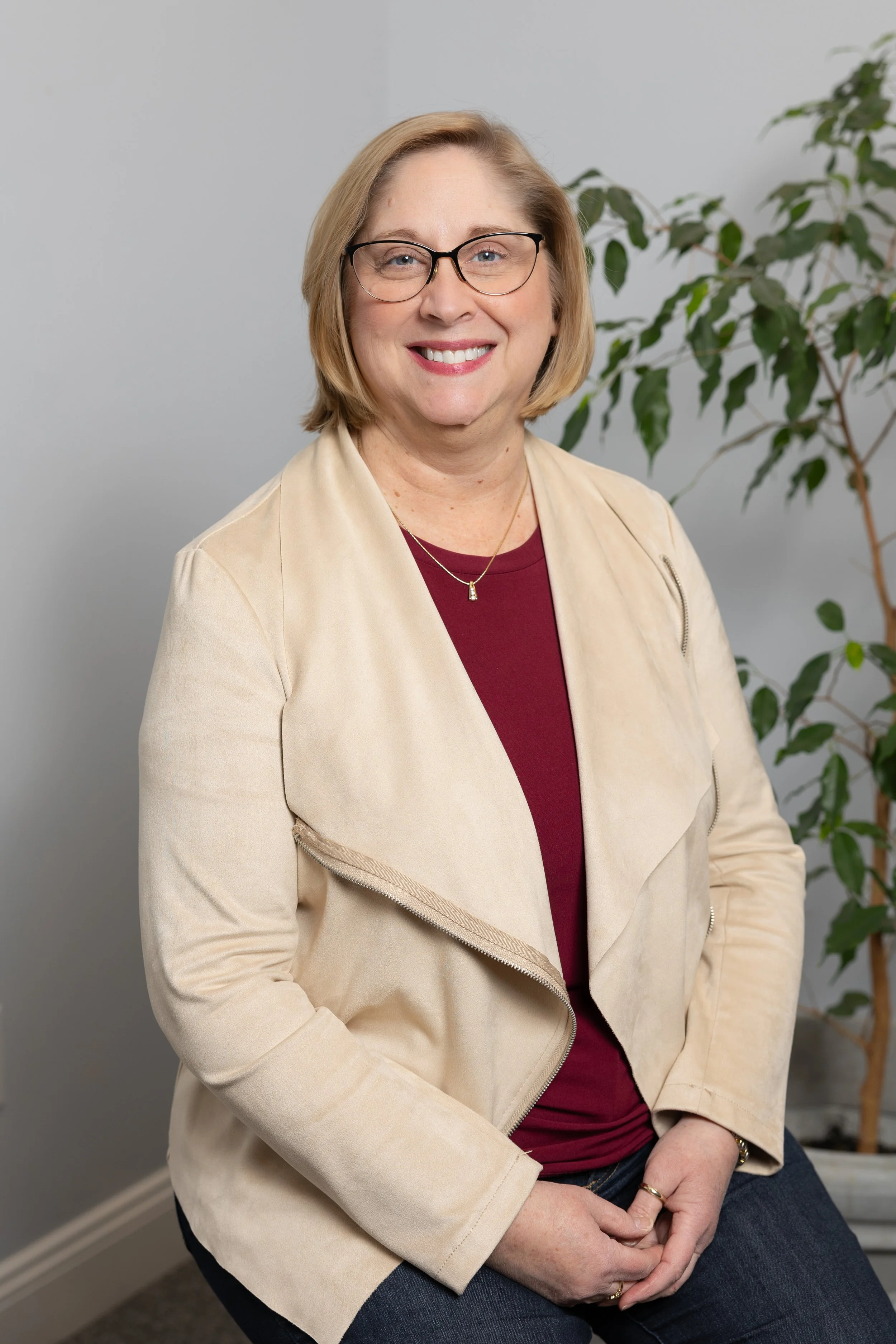 A woman with blonde hair, wearing glasses, a beige jacket, a red shirt, and dark jeans, sitting beside a green plant in a plain room.