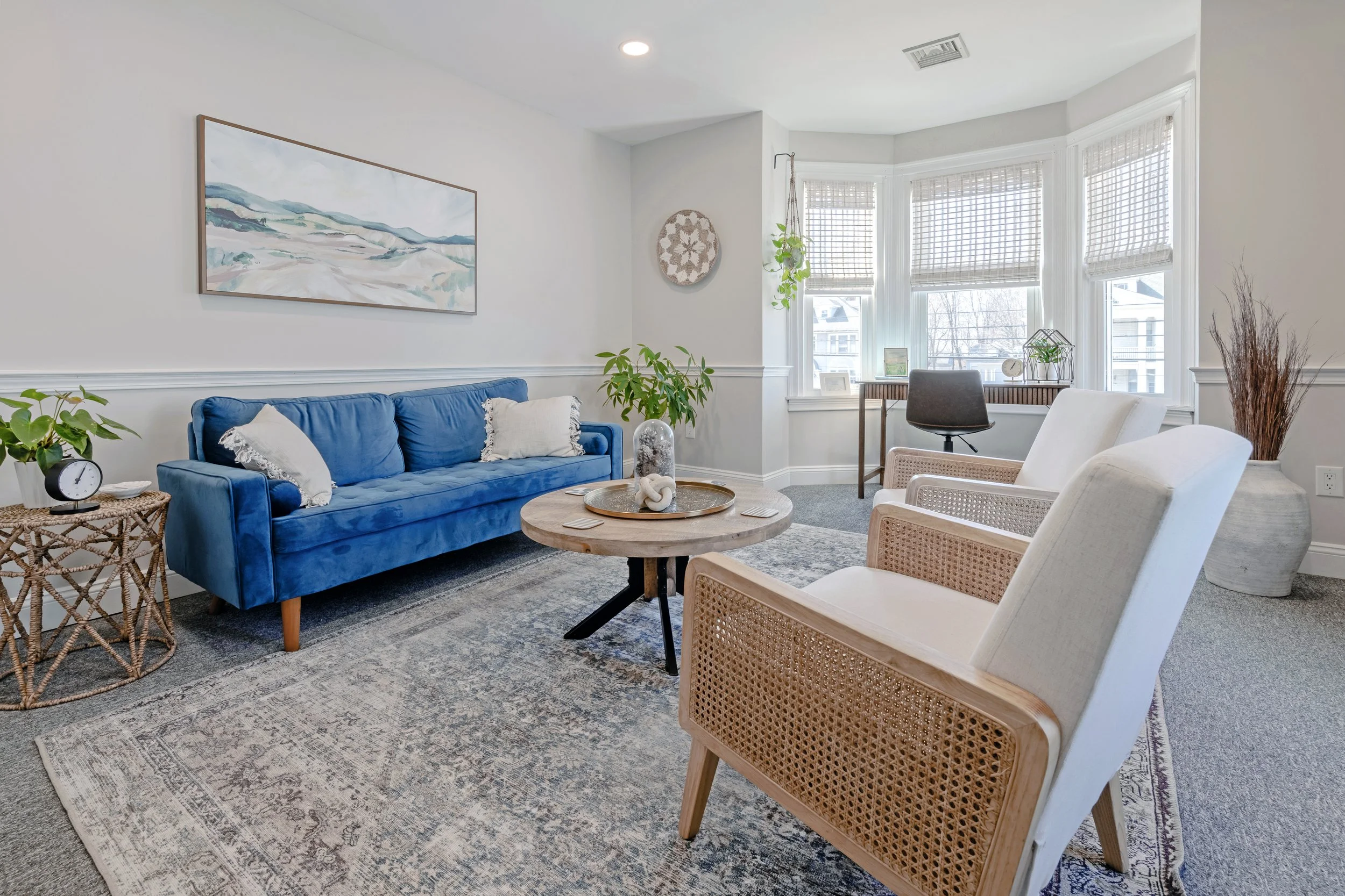 Room with a blue velvet sofa, white pillows, beige armchairs, a round coffee table, and a large window with blinds, decorated with plants and wall art.