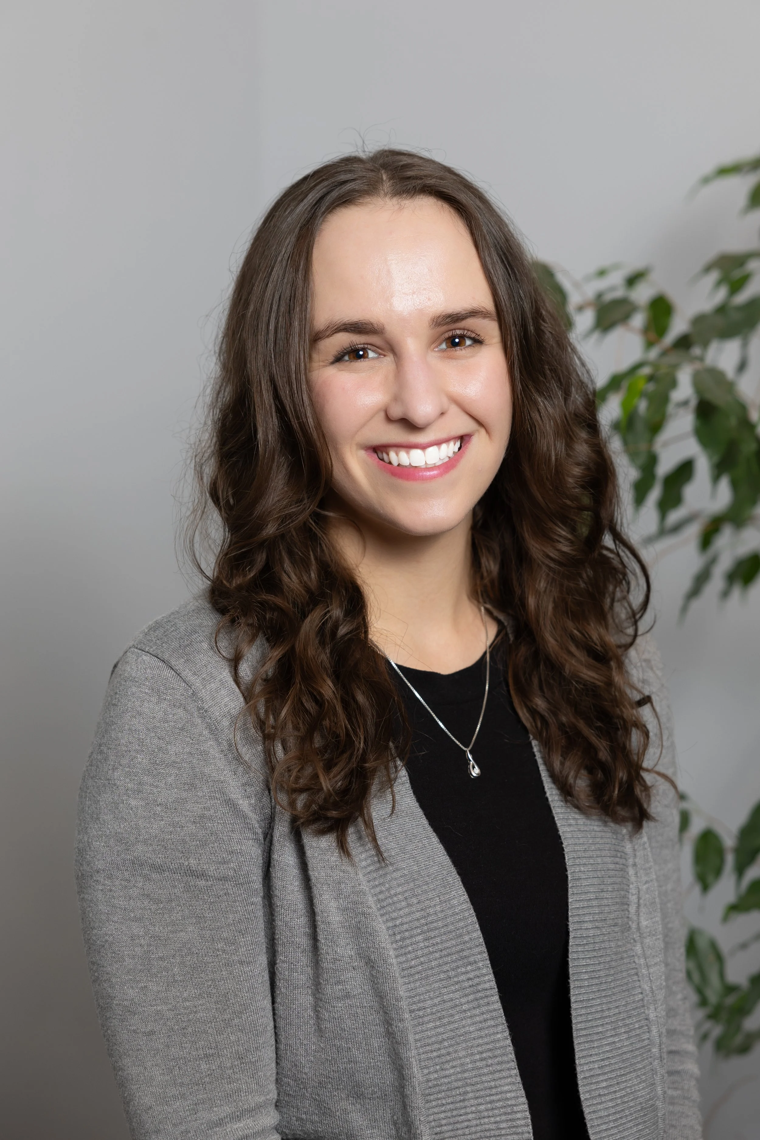A young woman with wavy brown hair, smiling, wearing a gray blazer over a black top, necklace, standing in front of a plain background with a green plant to her right.