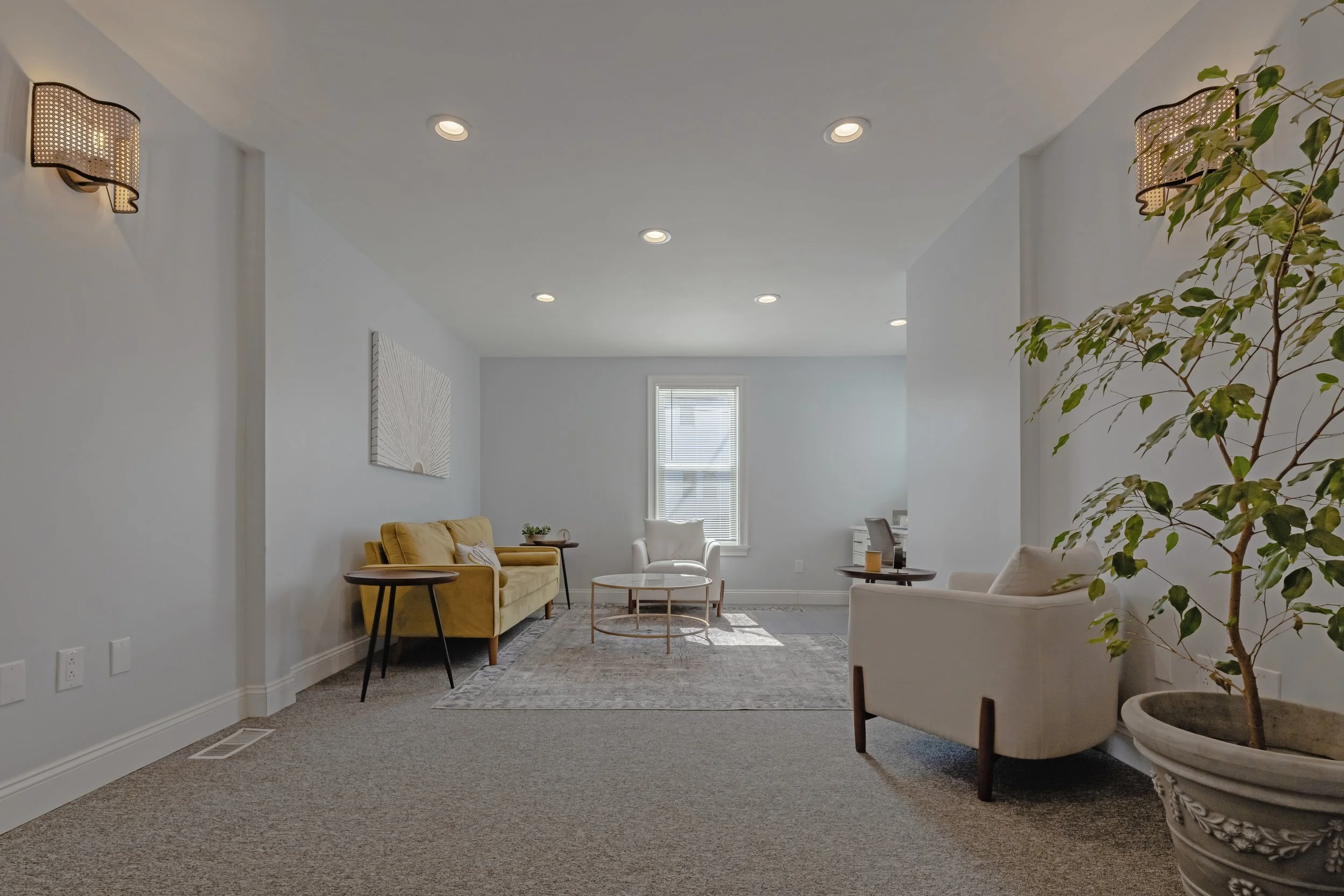 Room with white walls, carpeted floor, yellow sofa, white armchair, and a potted plant, illuminated by recessed ceiling lights and wall sconces, with a window letting in natural light.