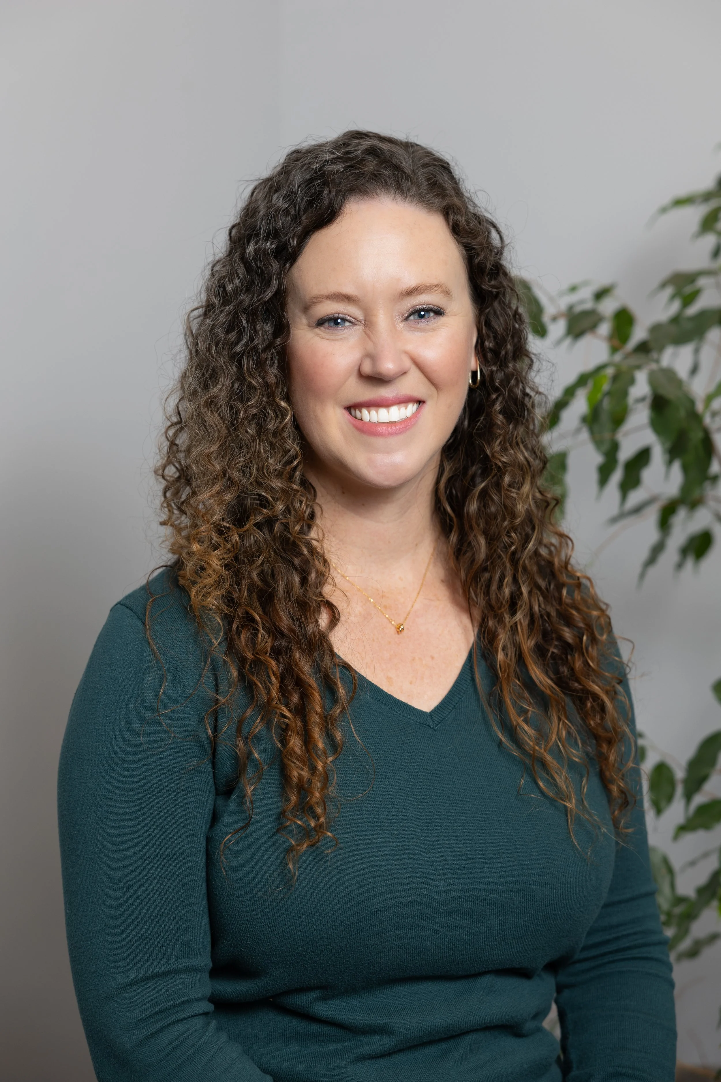 Portrait of a woman with curly brown hair, smiling, wearing a teal V-neck sweater and gold jewelry, standing in front of a plain wall and a green plant.