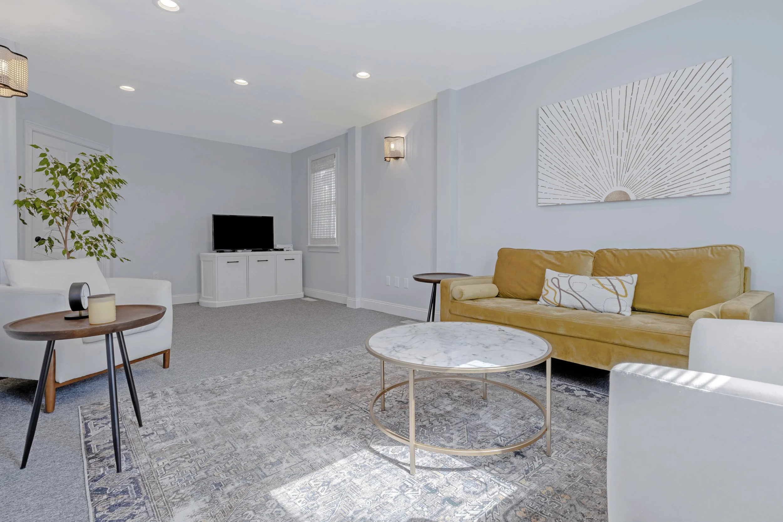 Room with light blue walls, gray carpet, and modern furniture including a yellow sofa, white armchair, marble coffee table, and wooden side tables. There is a flat-screen TV on a white cabinet, artwork on the wall, and a potted plant in the corner.