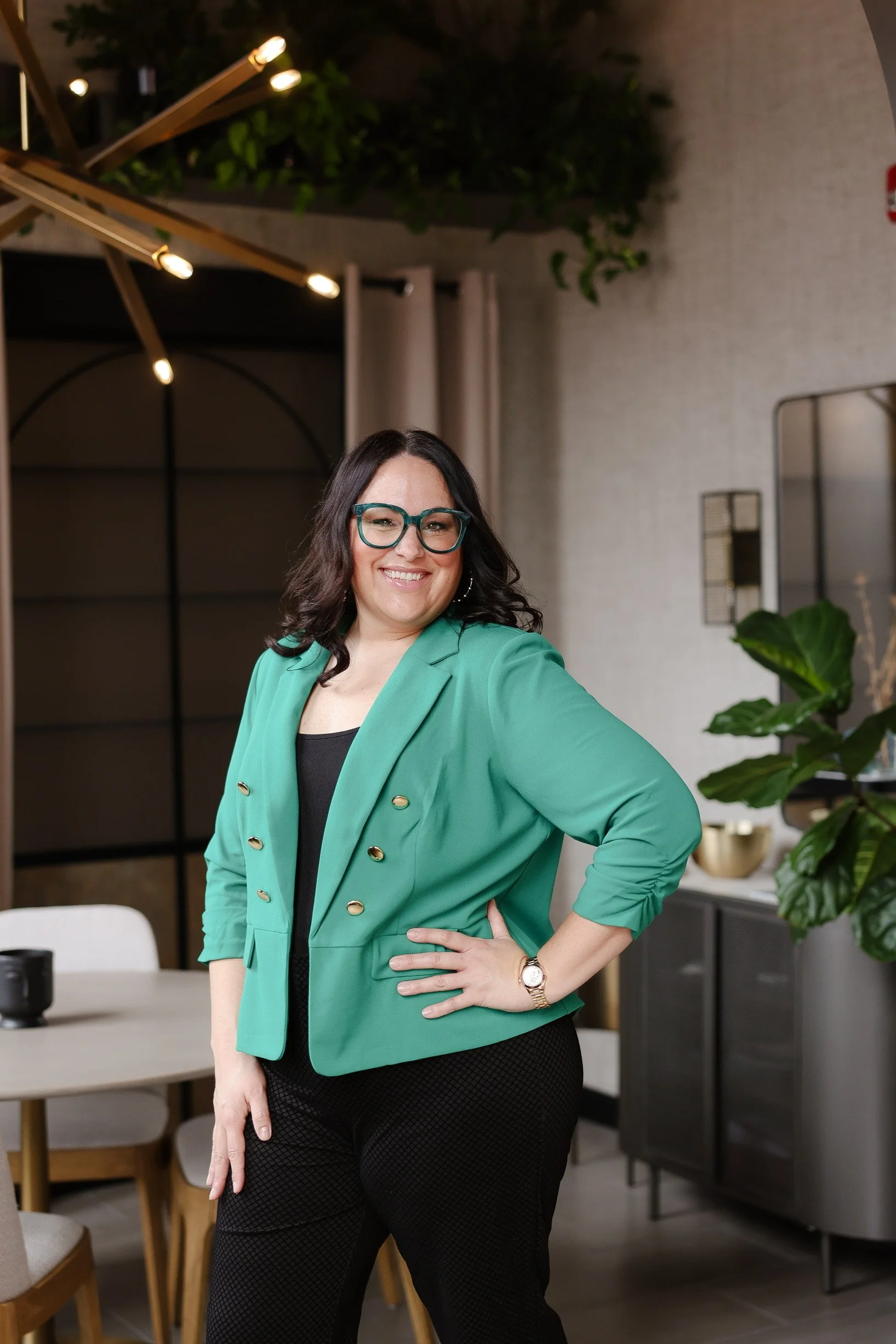 A woman with dark wavy hair, glasses, and a bright green blazer smiling and posing in a modern, stylish interior with plants and contemporary decor.