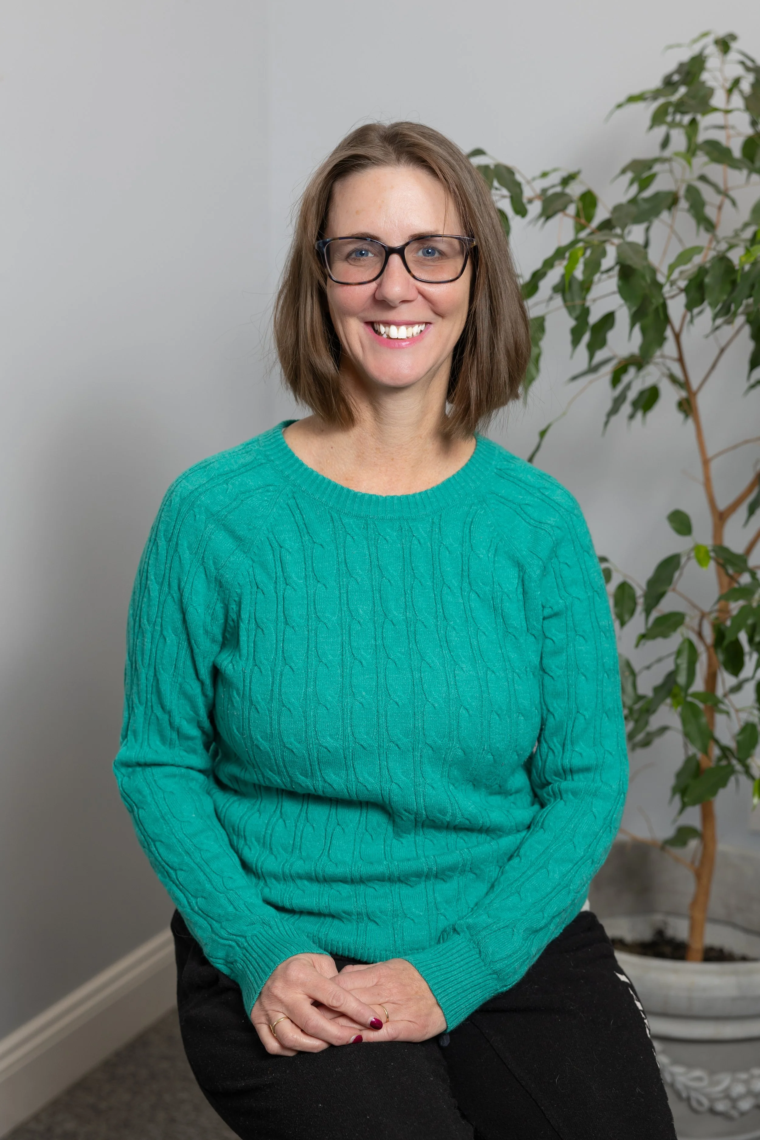 A woman with shoulder-length brown hair, wearing glasses and a teal sweater, smiling while sitting in front of a gray wall with a potted plant in the background.