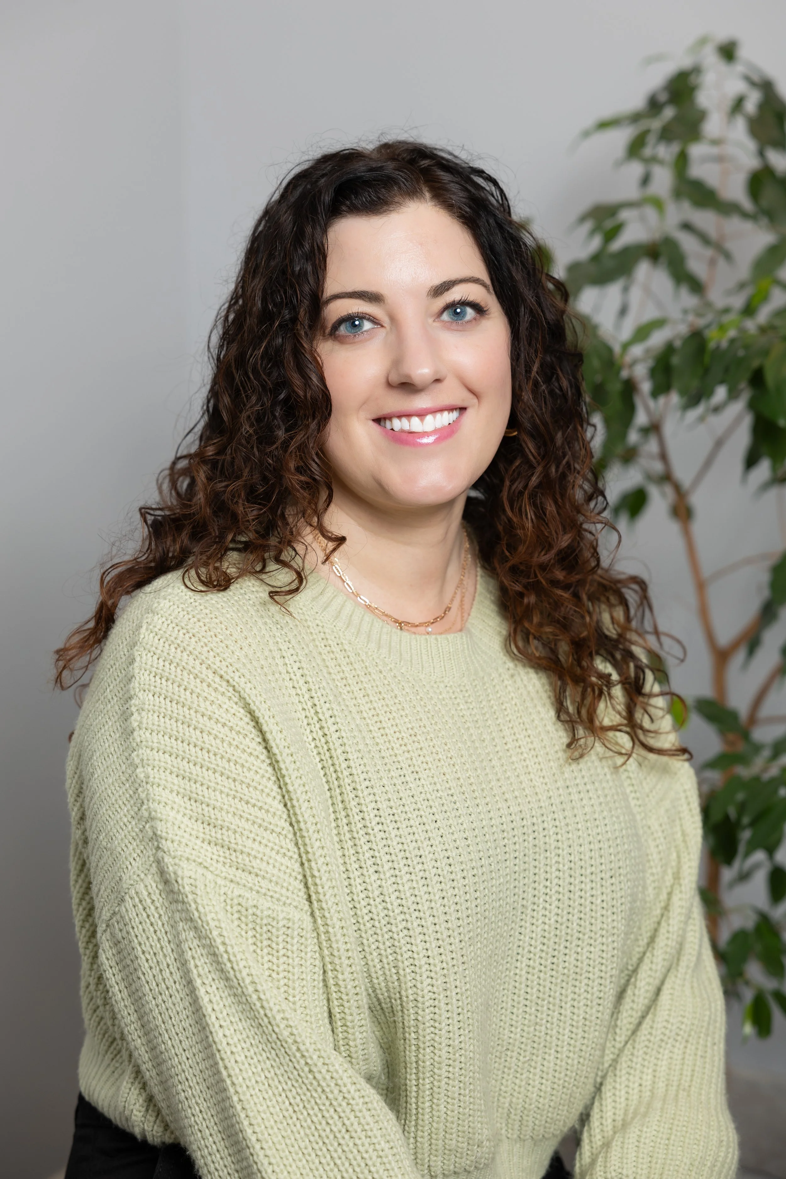 A woman with curly brown hair and blue eyes, wearing a light green sweater and gold jewelry, smiling in front of a gray wall and a green plant.