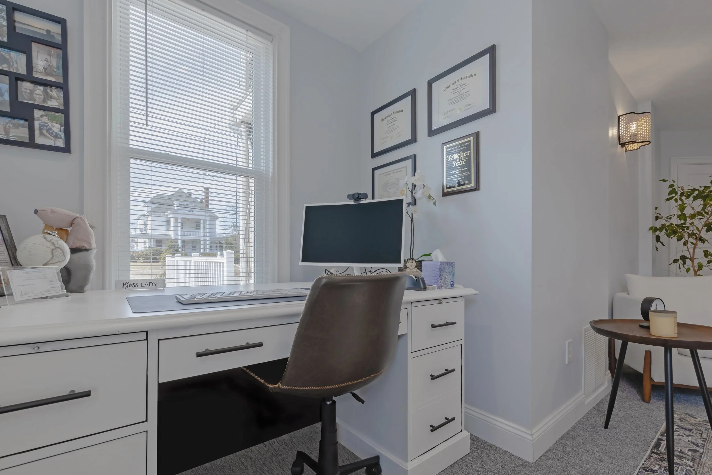 Office with white desk, computer monitor, framed certificates on wall, window with blinds, and a chair.