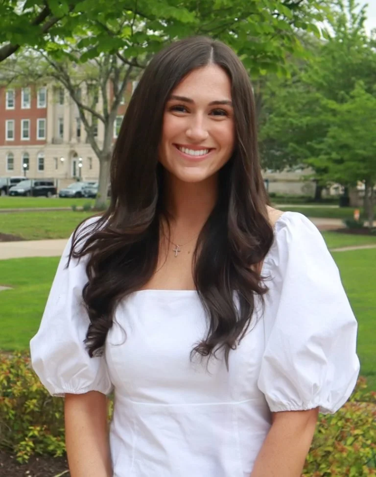A young woman with long dark hair wearing a white puff-sleeve top and a silver cross necklace, smiling outdoors in a park with green trees and a building in the background.