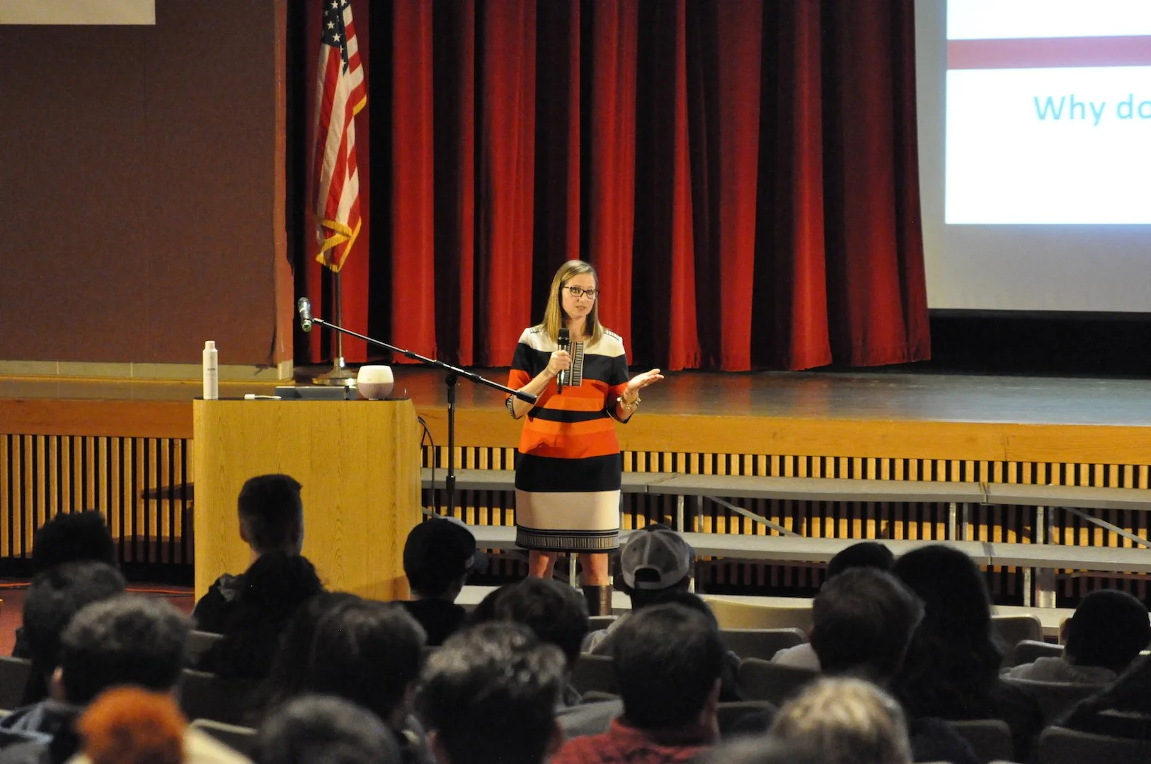 A woman giving a presentation on a stage with a microphone, in front of a large screen, with an audience seated in front. The stage has a red curtain and an American flag.