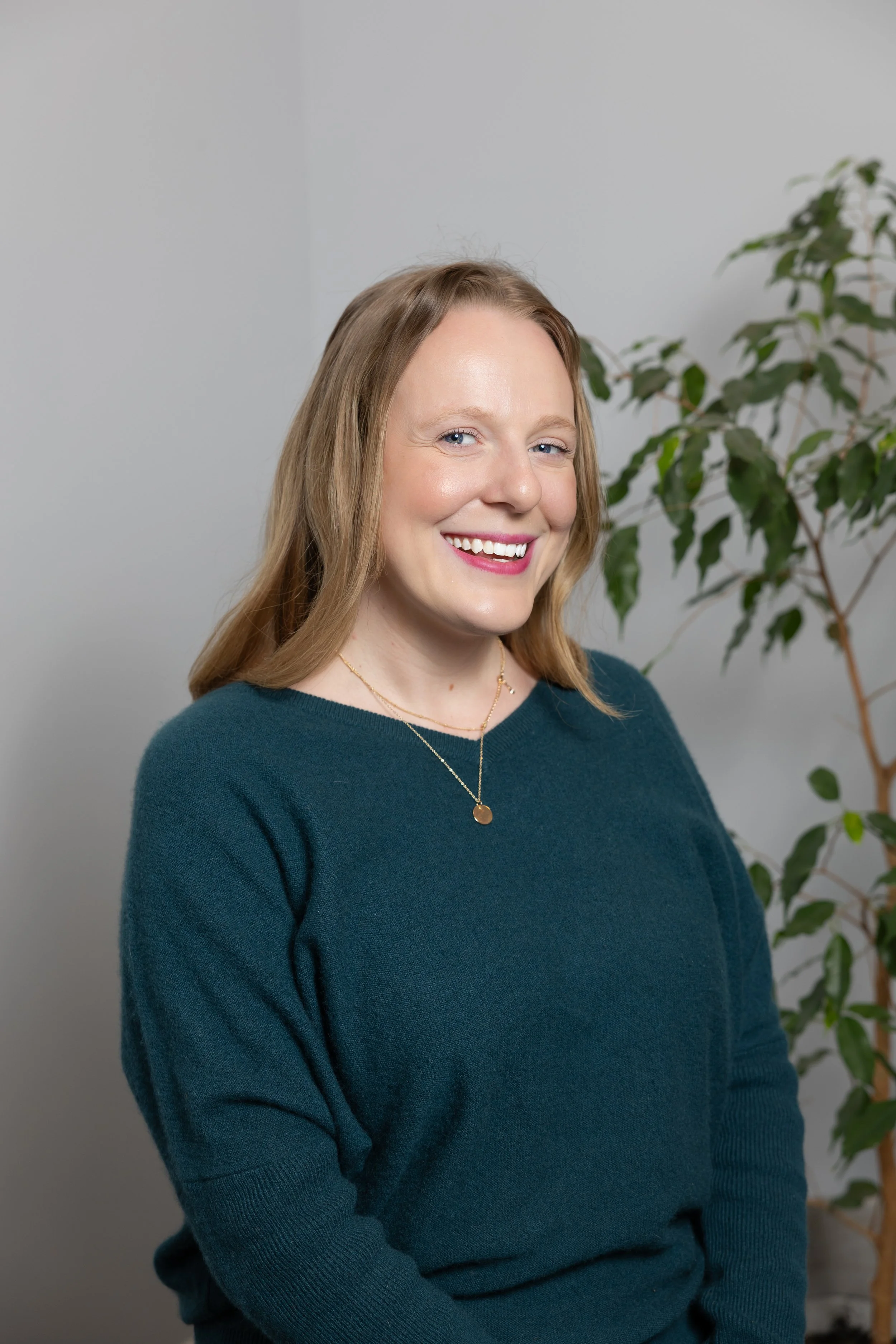 Portrait of a woman with blonde hair, wearing a teal sweater, smiling with a plant in the background.
