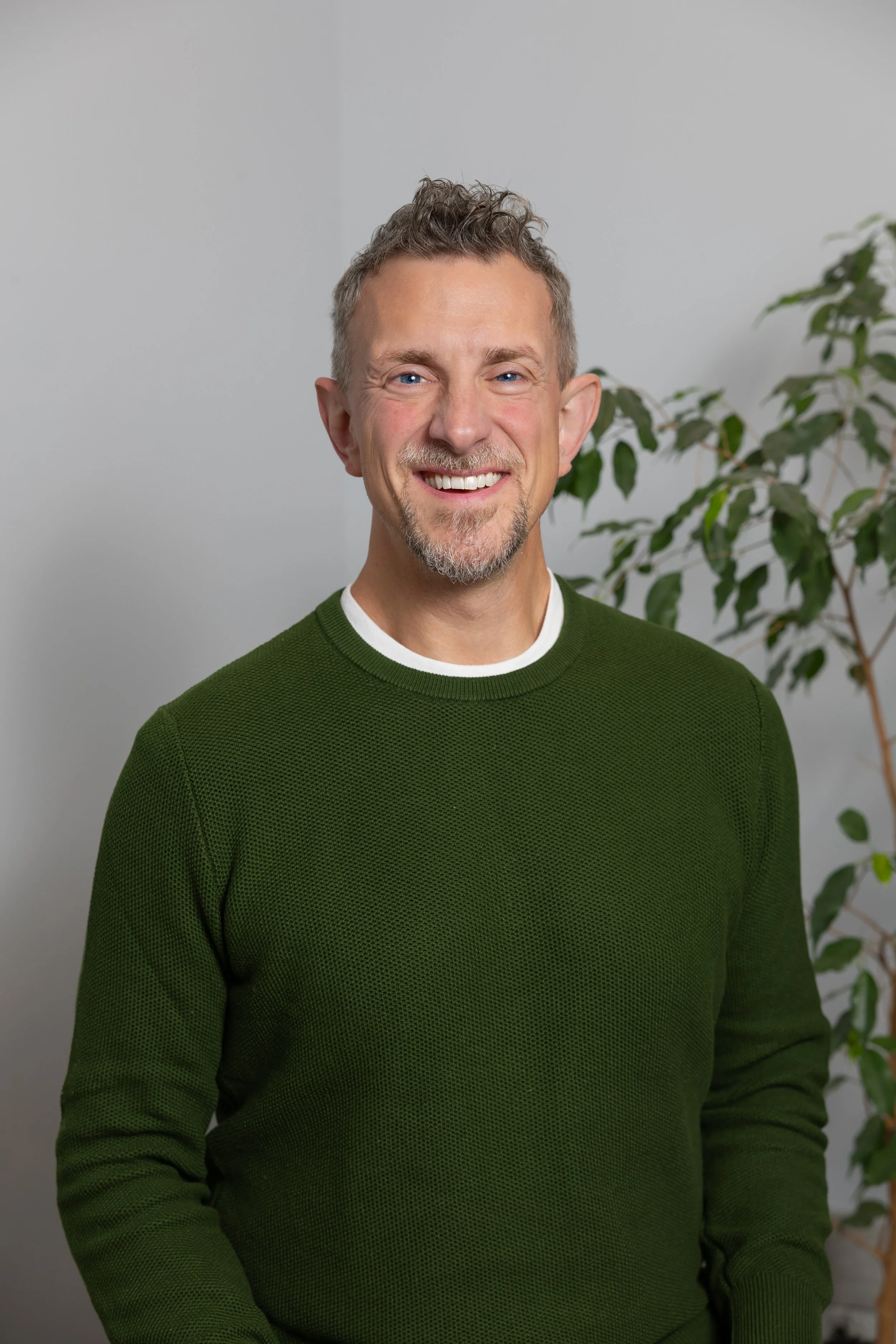 A smiling man with short curly gray hair, a beard, and blue eyes wearing a green sweater and standing in front of a light gray wall with a potted plant in the background.