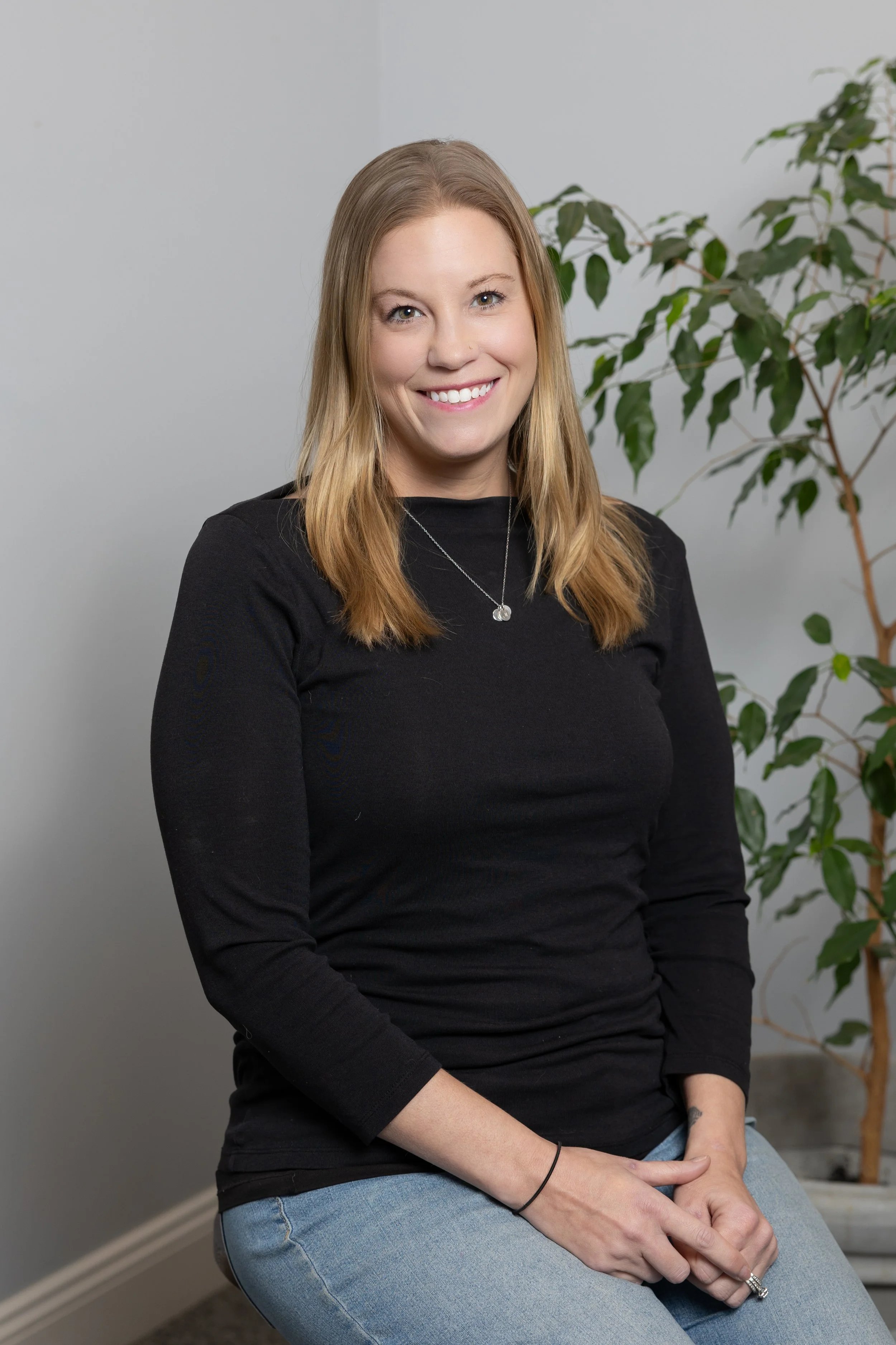 A woman with shoulder-length blonde hair smiling, sitting on a chair in a room with a gray wall and a potted plant in the background.