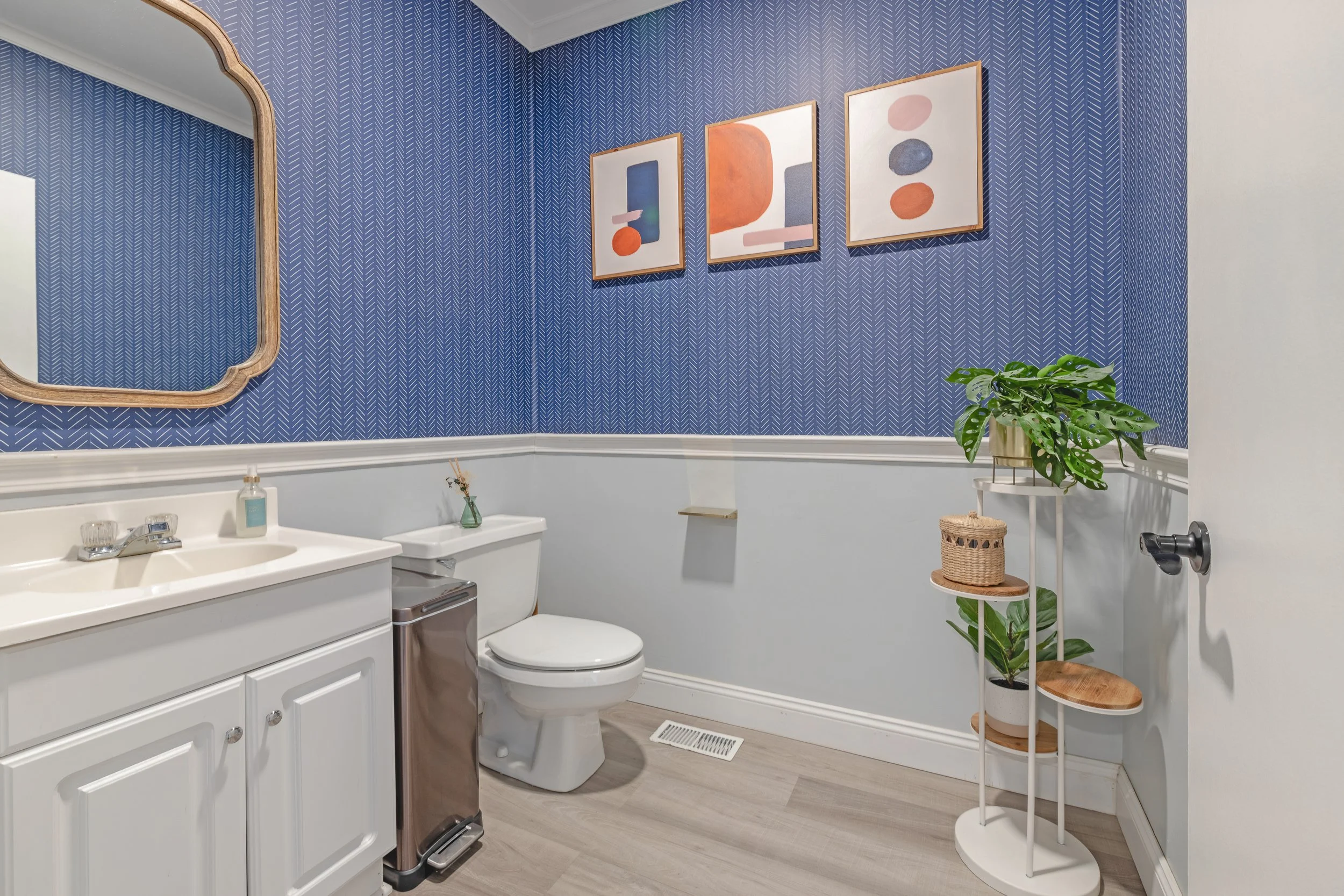 A small bathroom with a white vanity, a toilet, a mirror, and framed abstract art on a blue patterned wall. There are potted plants on a white stand and a small shelf above the toilet.