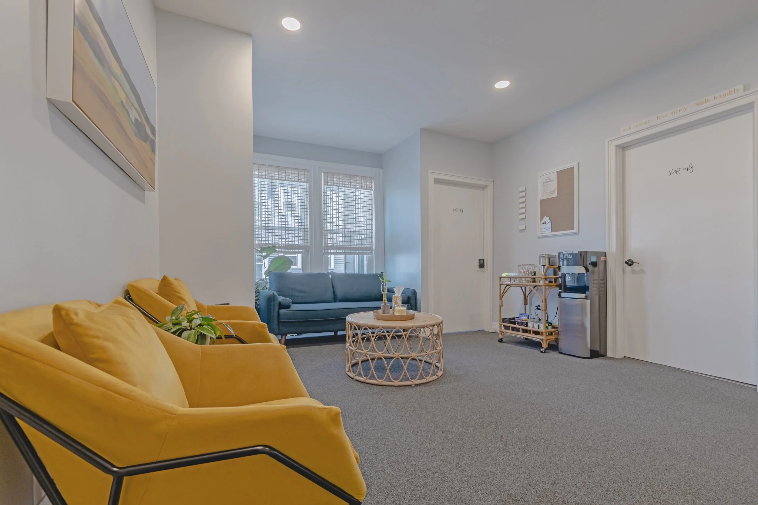 A waiting room with yellow couches, a blue armchair, a round rattan coffee table, windows with bamboo shades, a cart with drinks and glasses, a small water dispenser, and framed artwork on white walls.