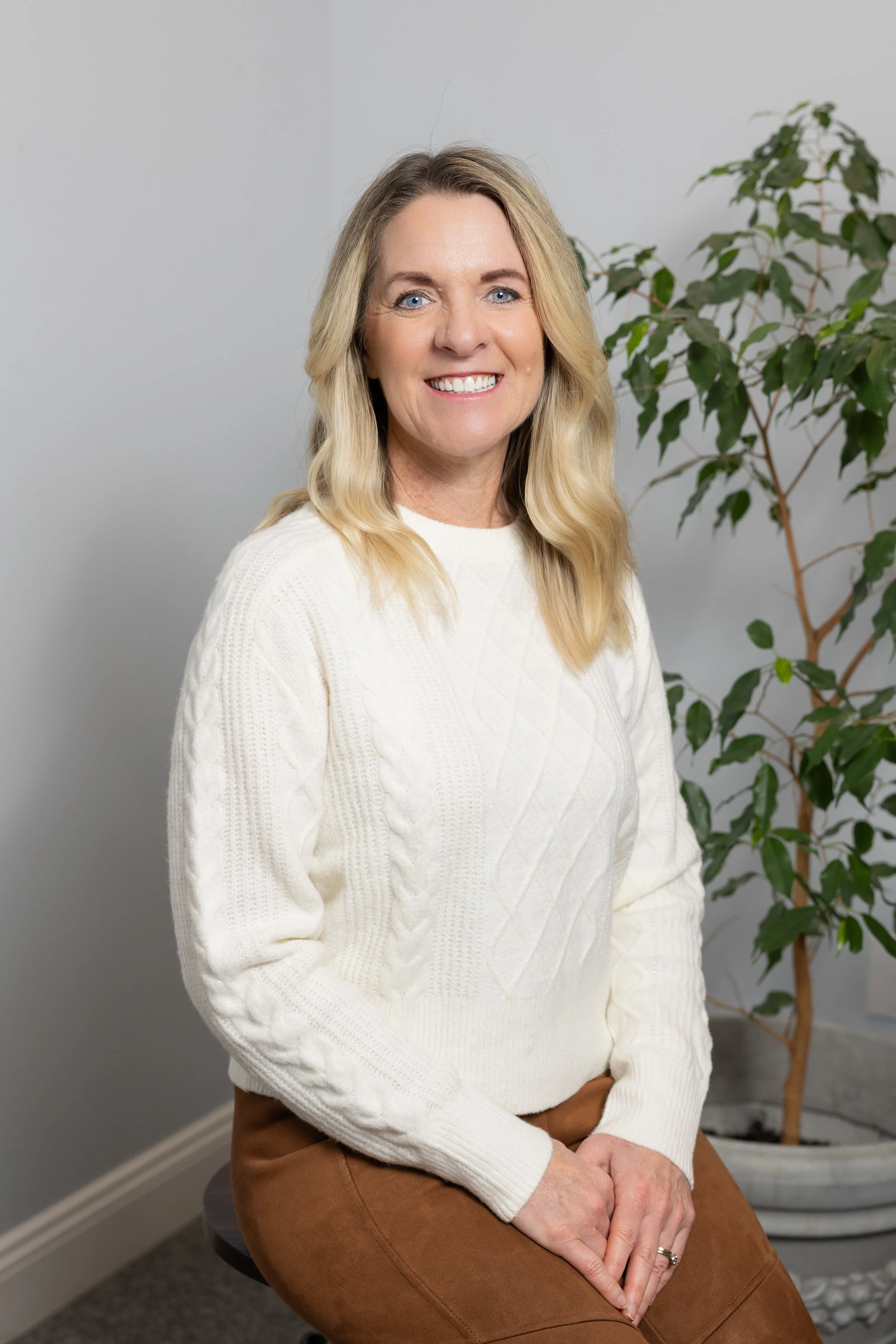 A woman with blonde hair, wearing a white sweater and brown pants, sitting on a stool indoors with a plant in the background, smiling at the camera.