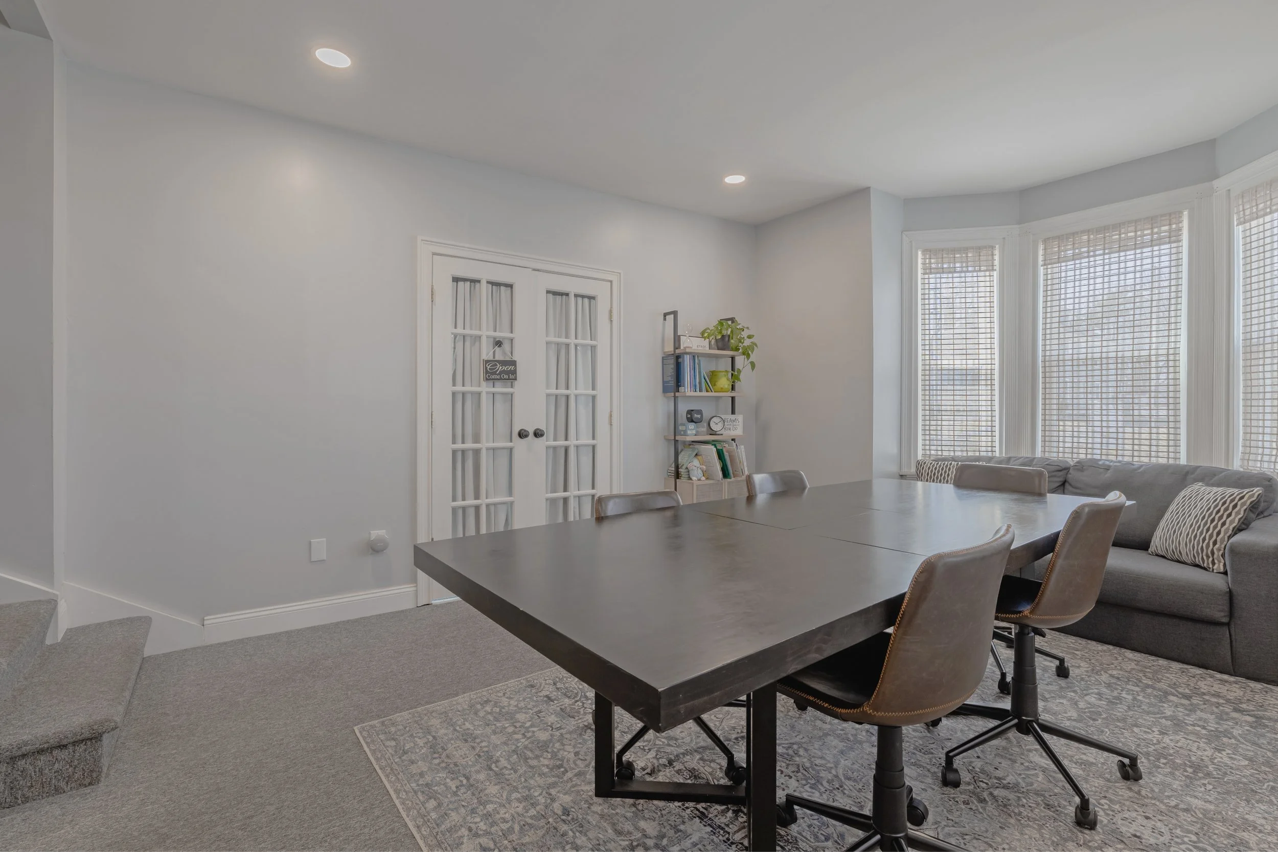 A bright office or meeting room with a large dark wooden table, cushioned office chairs, and a gray sofa with pillows, near windows with blinds, and a small bookshelf with plants and books.