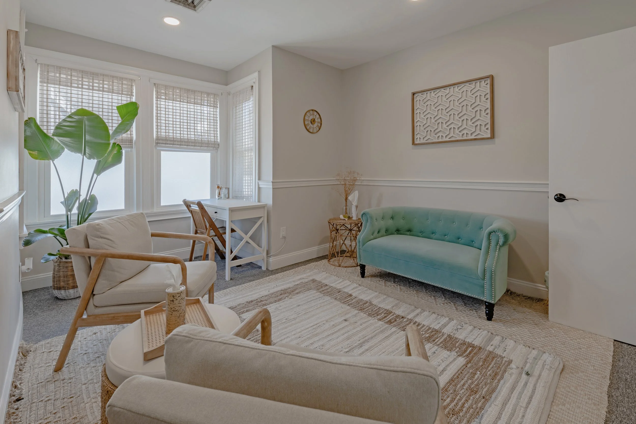 Room with cream-colored sofas, a green velvet settee, a wicker side table with dried flowers, a white desk near a bay window with woven blinds, and a large houseplant; decorated with minimal wall art and a neutral area rug.
