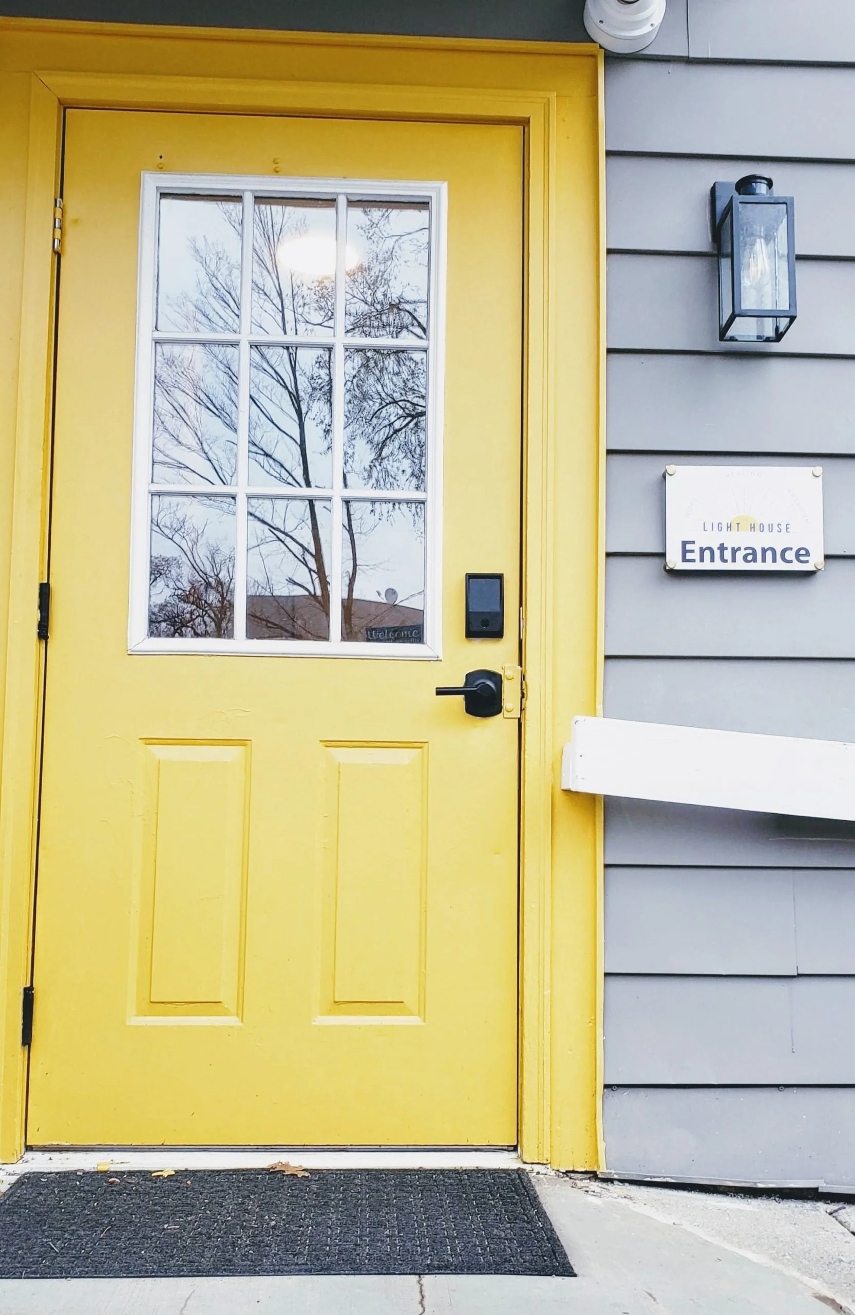 Yellow door with a window and an electronic lock, part of a gray building with a sign that reads 'LIGHT HOUSE Entrance', an outdoor wall lantern, and a black doorbell.