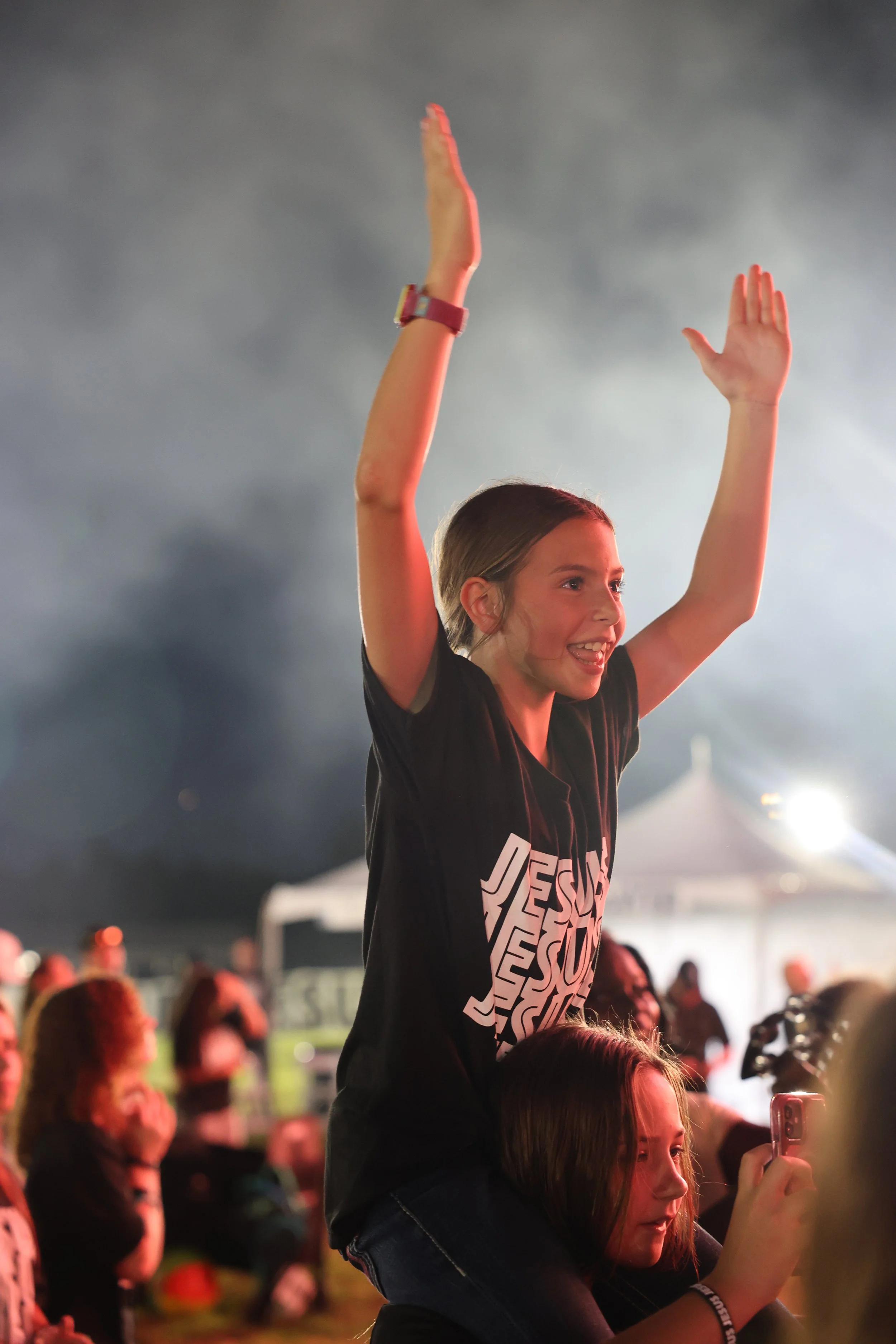 Young girl with long hair sitting on another person's shoulders at an outdoor event, with her arms raised and smiling, surrounded by a crowd at night.