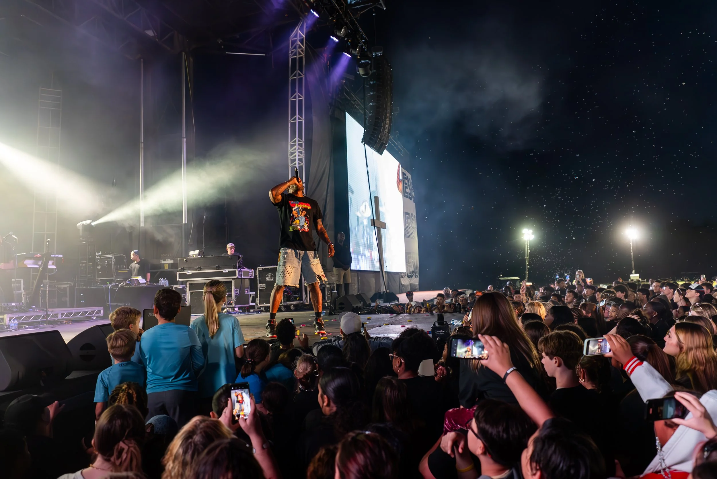 Singer performing on an outdoor stage at night, with a large crowd of fans, some taking photos, and bright stage lights under a starry sky.