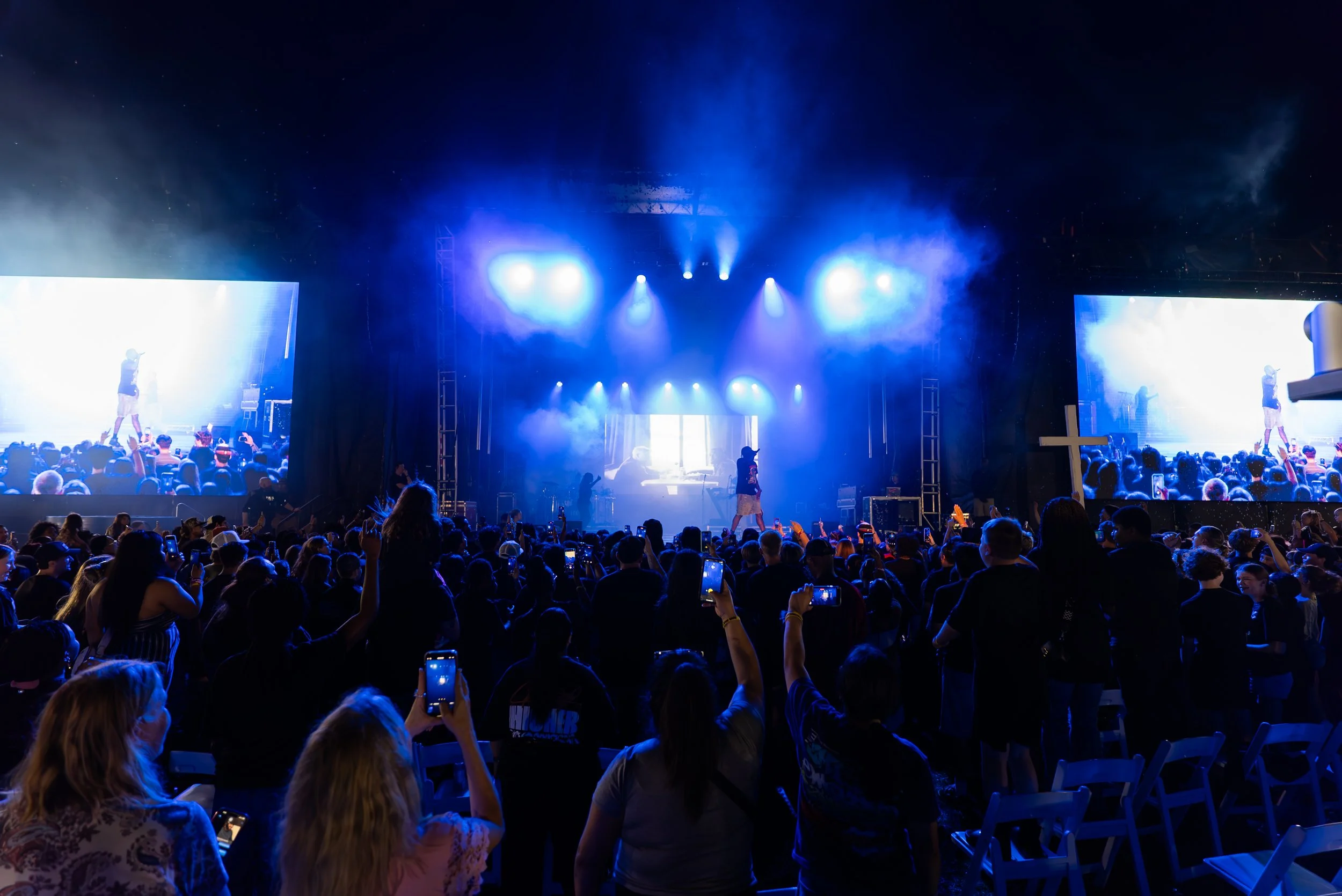 Crowd watching a concert on stage with large screens and blue lighting at night.