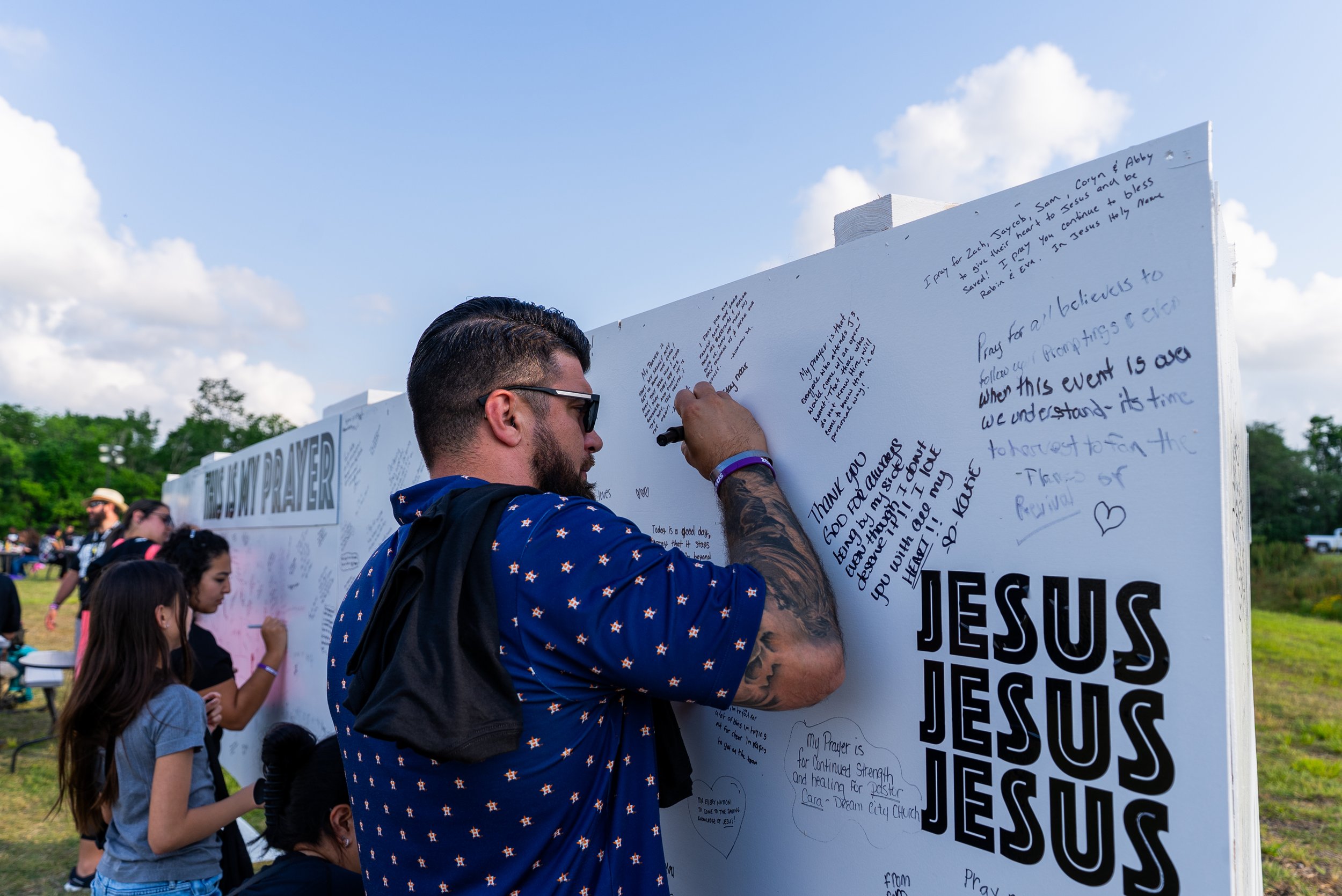Man with sunglasses and tattoos writing on a large white prayer board outdoors, with children and other people in the background, some of whom are also writing on the board.