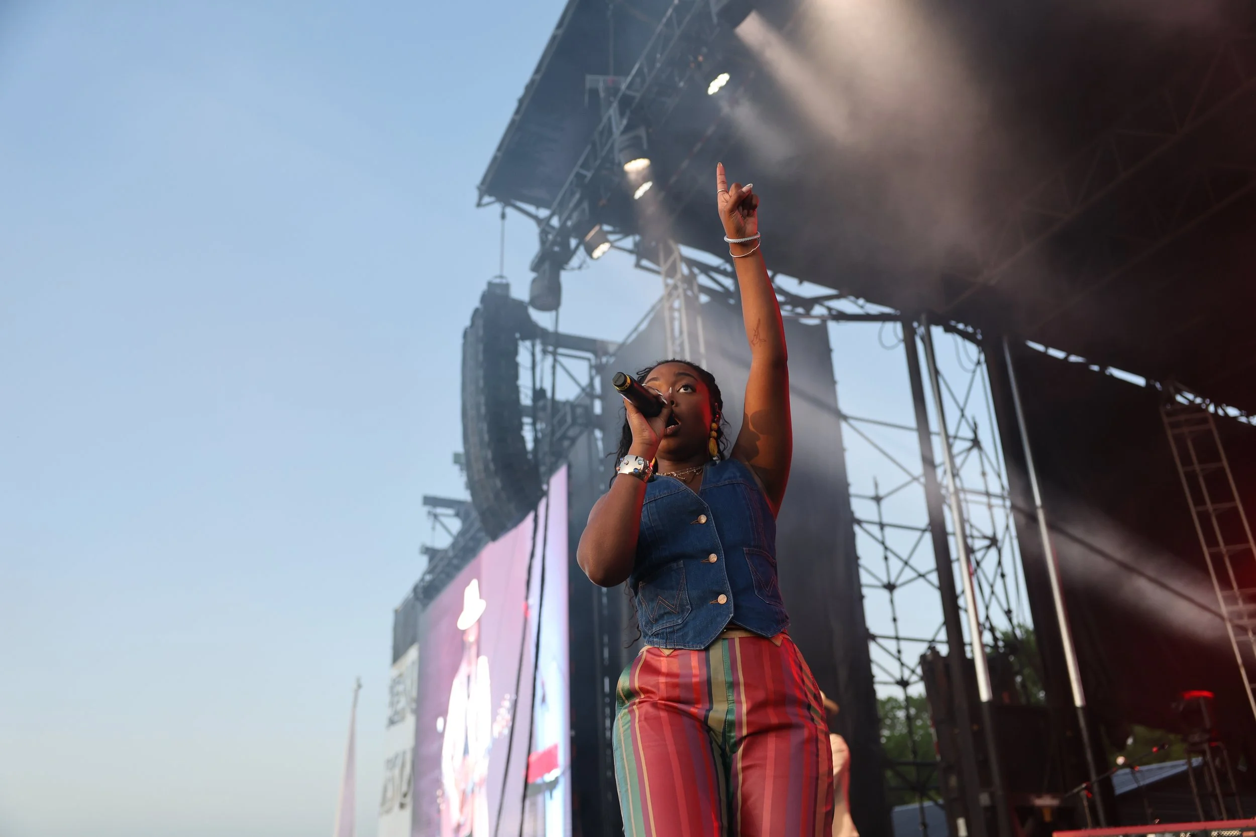 Performer singing on stage during outdoor concert, pointing upwards with her right hand, wearing colorful striped pants and a denim vest, stage lights and equipment visible in the background.
