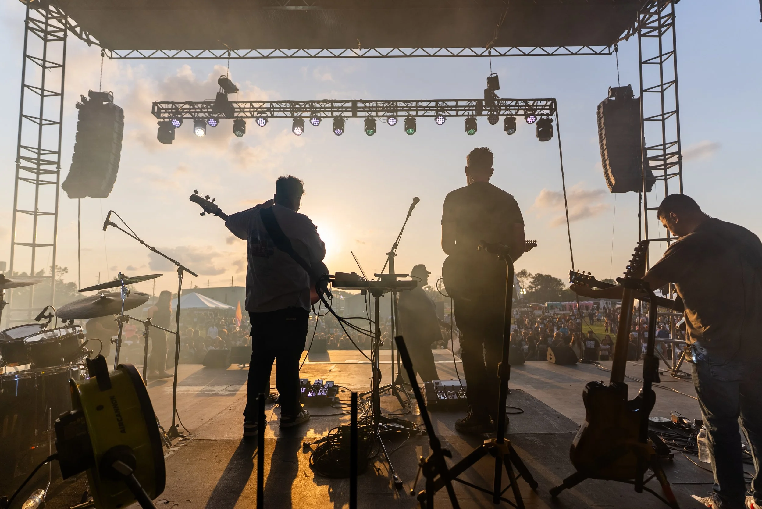 Musicians playing instruments on an outdoor stage at sunset during a live performance, with a crowd of people in the background.
